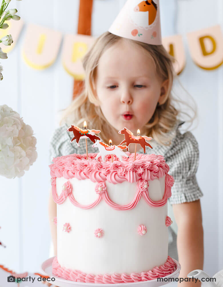A preschool aged girl in her green gingham dress, wearing Momo Party's floral pony birthday party hat and blowing Momo Party's floral pony candles by Party Deco on a pink and white vintage buttercream cake. Behind her is a blush happy birthday banner hung on the wall. An adorable pony horse themed birthday party set up for a little girl.