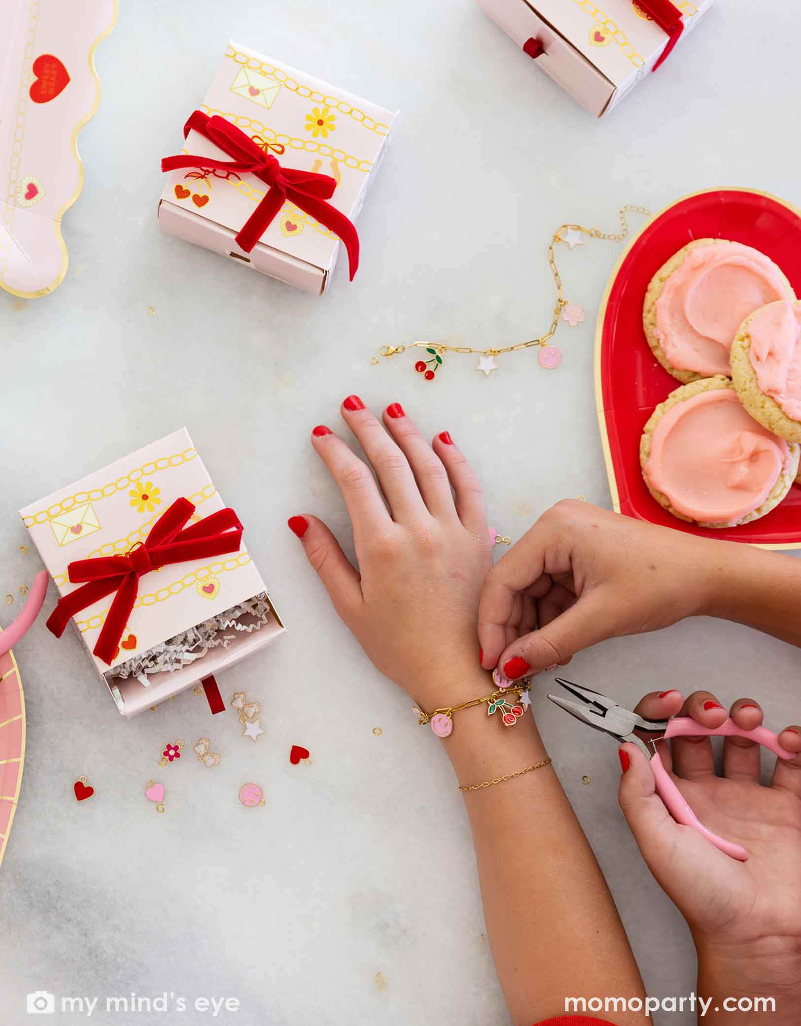 Children's hands with pink nail polish working on their charm bracelets with pink pliers, surrounded by small charms, gift boxes and Momo Party's Valentine's Day charm matching party tablewares including heart shaped plates and napkins on the table. 