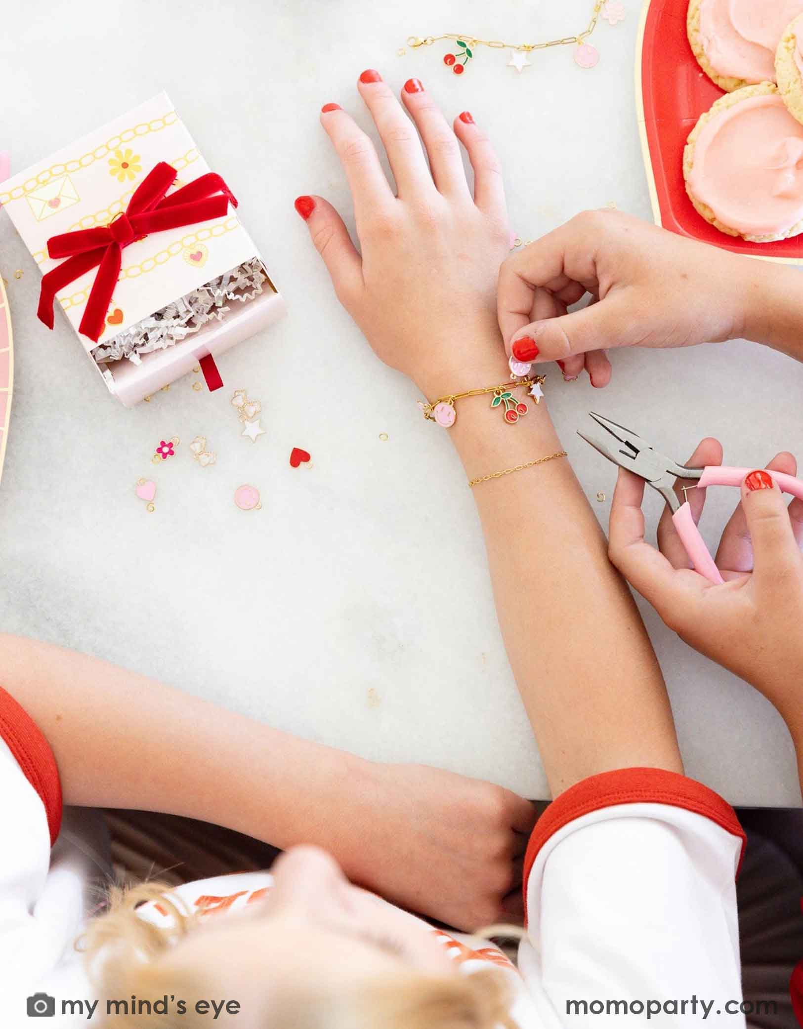 Children's hands with red nail polish working on their charm bracelets with pink pliers, surrounded by small charms, gift boxes and Momo Party's Valentine's Day charm matching party tablewares including heart shaped plates and napkins on the table. 