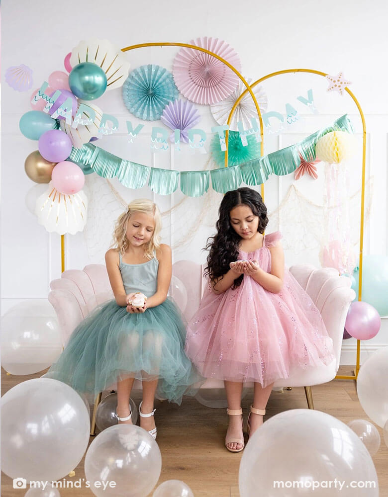Two school-aged girls sitting on a shell shaped blush sofa in an enchanting kid's mermaid birthday party set up featuring mermaid inspired decorations and balloons from Momo Party including the Under the Sea paper fan set in pastel colors of light pink, aqua, teal, lilac and cream, Mermaid Happy Birthday Banner Set with aqua foil fringe banner and shell shaped paper decorations by My Mind's Eye on the wall. On the floor are some clear latex balloons in various sizes creating a whimsy water bubble scene.