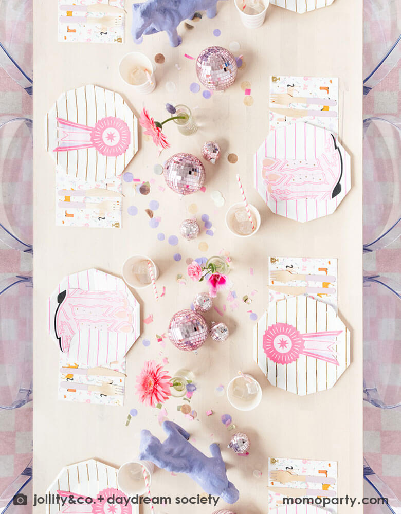 A table shot from the top of a girl's rodeo pony themed party table decoration features Momo Party's cowgirl themed party supplies by Daydream Society, including cowgirl boots shaped napkins, pink striped party plates, cups and cowgirl motif large napkins. In the middle of the party table, there are two purple pony figures as the centerpiece with some pink disco balls, making this an adorable inspiration for girl's First Rodeo first birthday celebration or a cowgirl theme party for pony-loving girls!