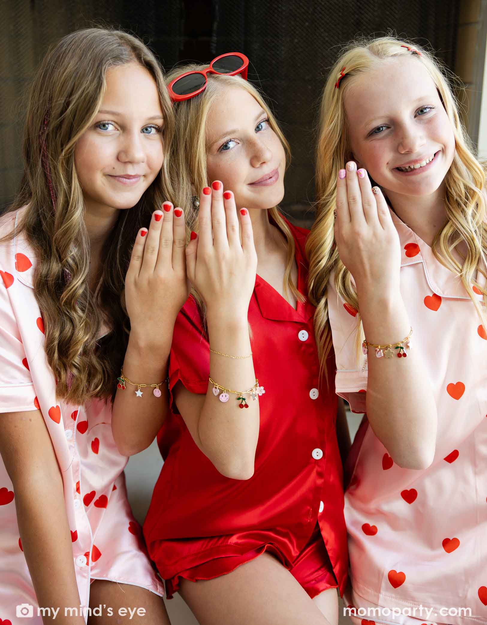 Three young girls in Valentine's Day pajamas with heart patterns showing their hands with red and pink polish and the charm bracelets from Momo Party posing together.