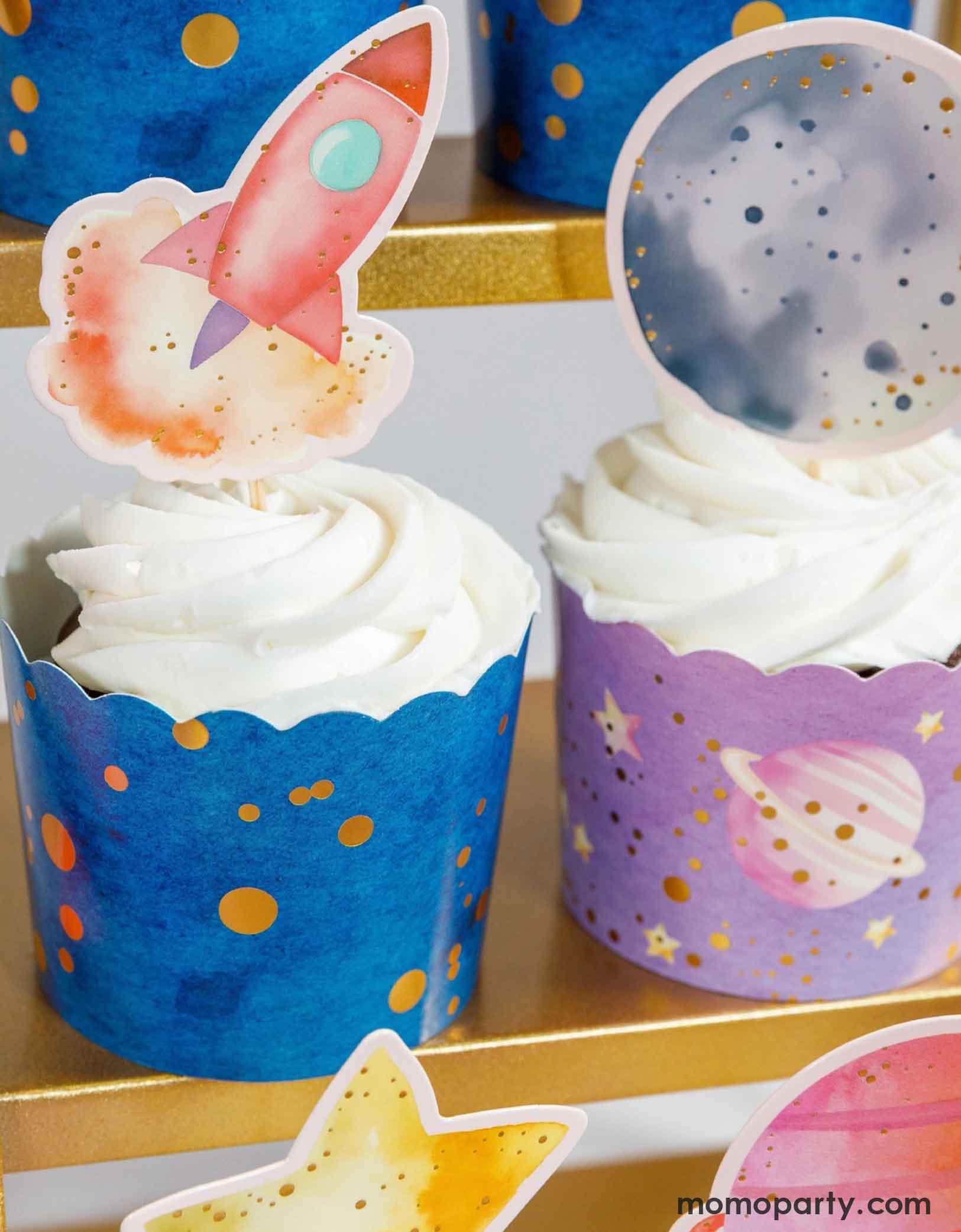 Cupcakes with Momo Party's space-themed toppers including a pink rocket ship and a moon and colorful wrappers on a gold cupcake stand.