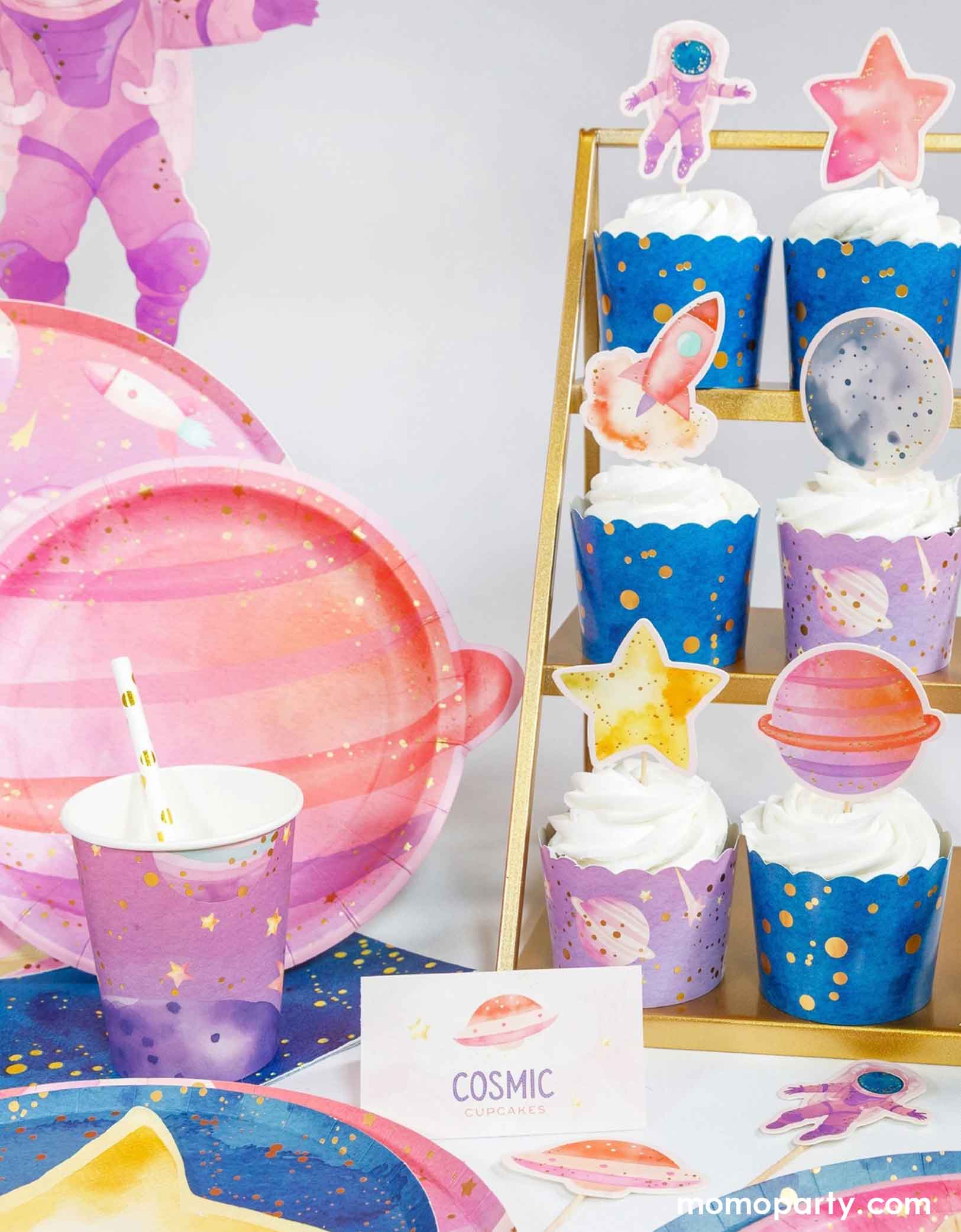 A girl's space theme party table set up featuring a cupcake stand with cupcakes wrapped in lilac and galaxy blue wrappers and topped with Momo Party's space-themed cupcake toppers including a pink astronaut, a pink rocket ship, planets and a star. Next to is are various pastel colored space-themed party supplies including cups, plates, napkins with cosmic decorations.