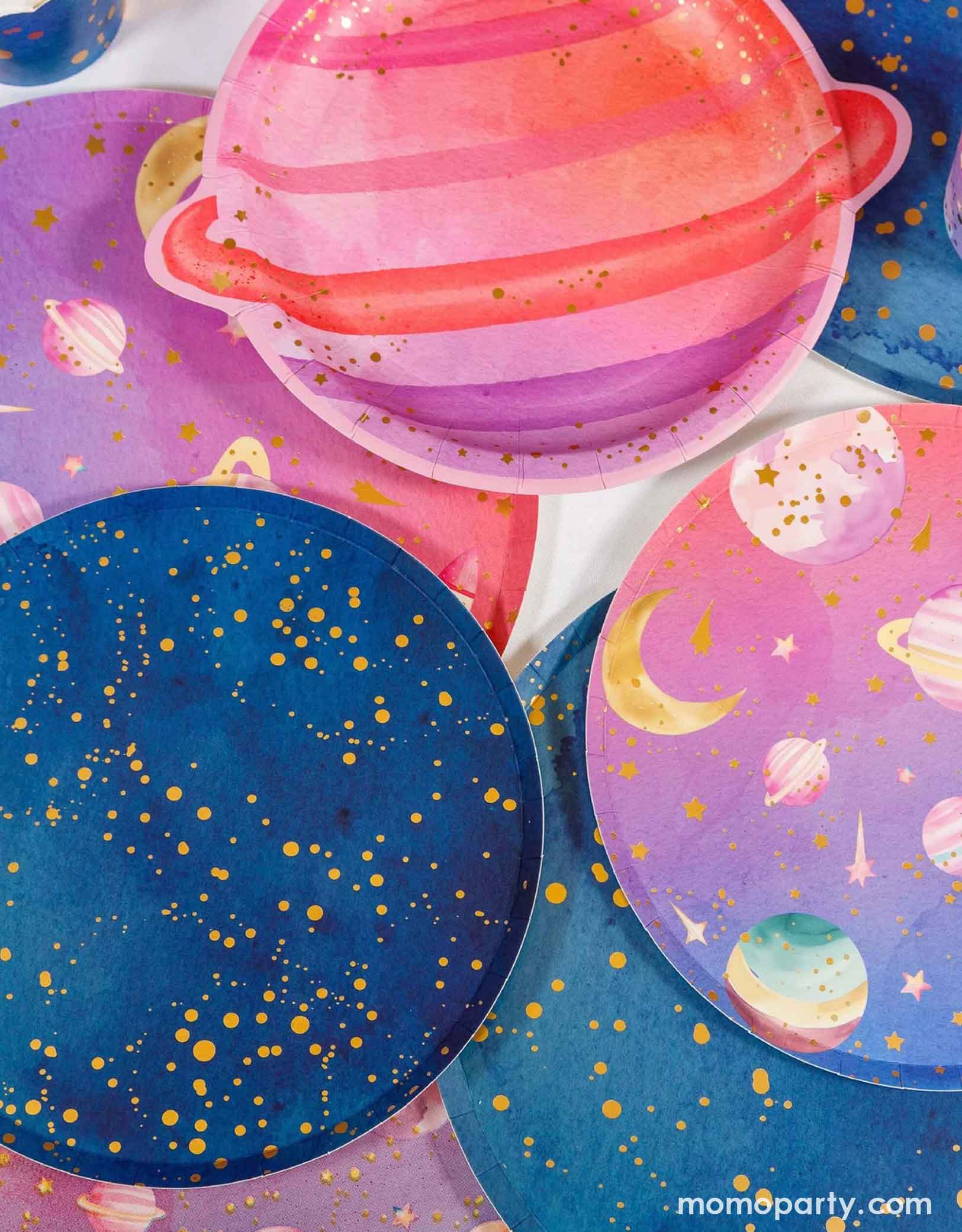 Various space themed party plates from Momo Party on a table including a pink planet shaped plate, blue galaxy plates with gold dots on them, and pink and lilac ombre space plates.
