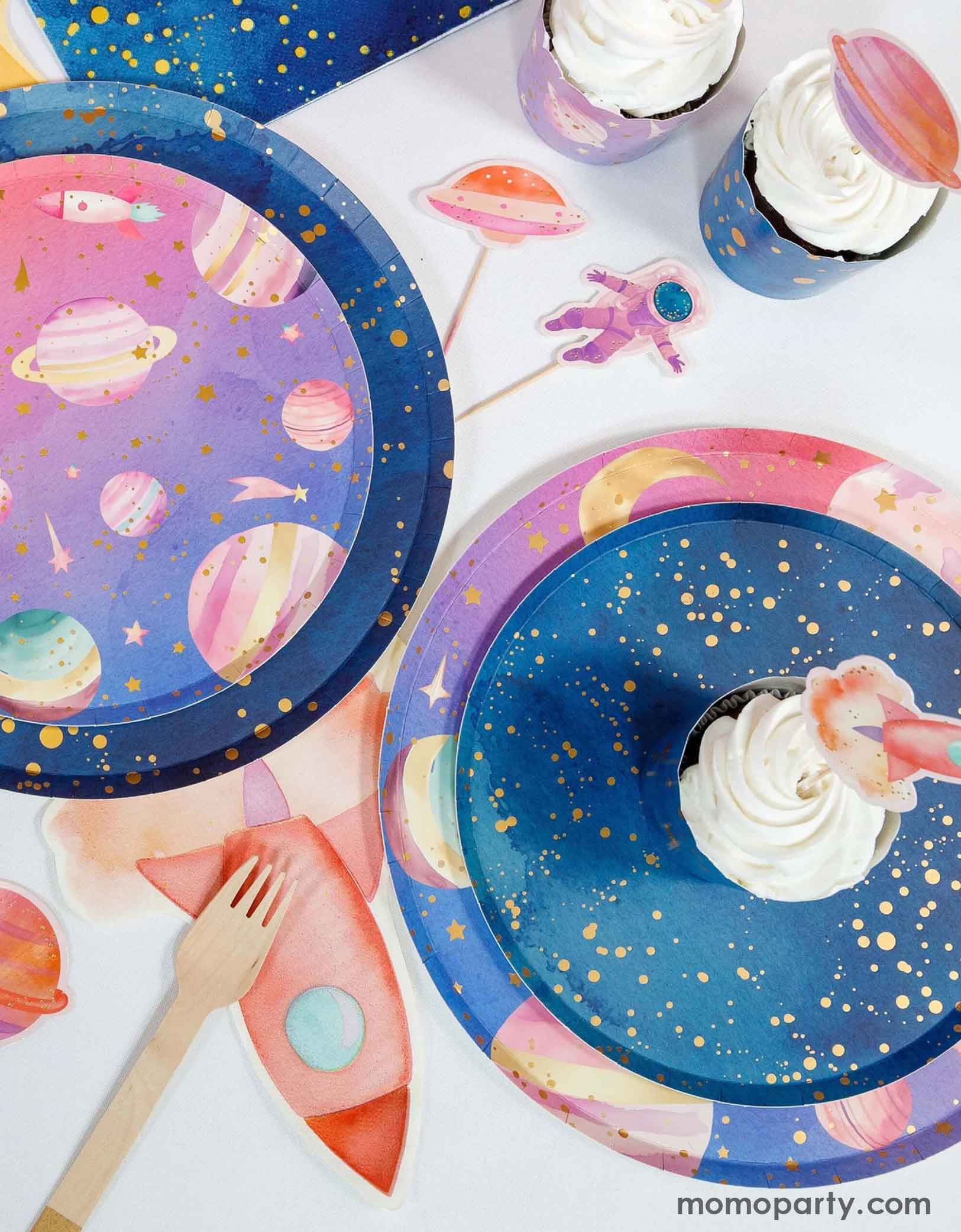 Various girly space themed party tablewares from Momo Party on a party table, including party plates, rocket ship shaped napkins cupcakes with space themed toppers on a white background.