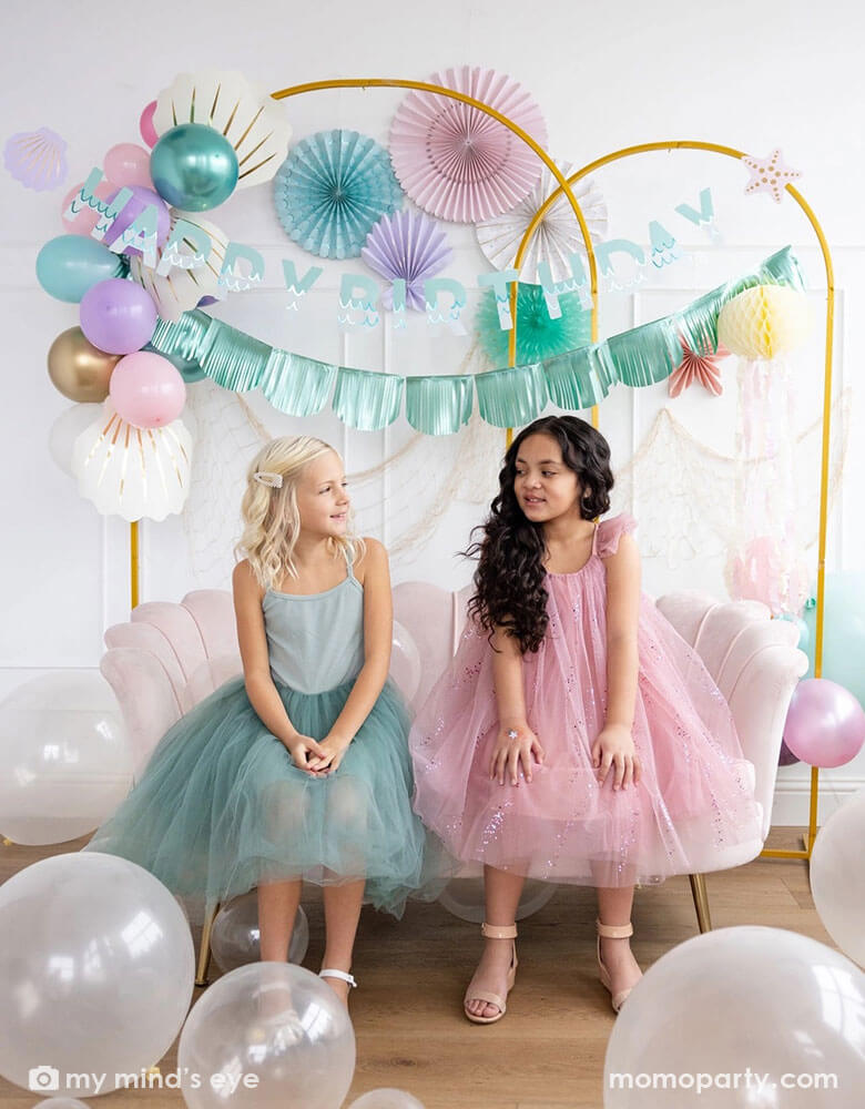 Two school-aged girls sitting on a shell shaped blush sofa in an enchanting kid's mermaid birthday party set up featuring mermaid inspired decorations and balloons from Momo Party including the Under the Sea paper fan set in pastel colors of light pink, aqua, teal, lilac and cream, Mermaid Happy Birthday Banner Set with aqua foil fringe banner and shell shaped paper decorations by My Mind's Eye on the wall. On the floor are some clear latex balloons in various sizes creating a whimsy water bubble scene.