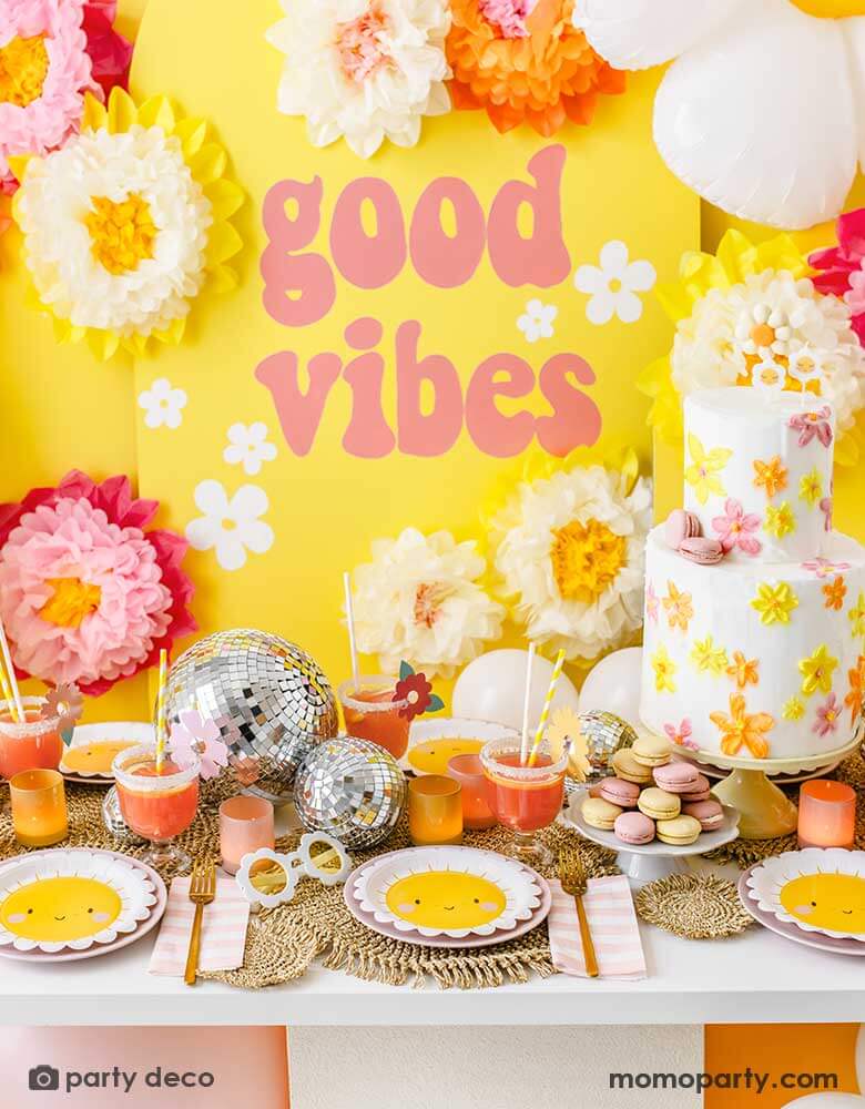 A "Good Vibes" themed party featuring Momo Party's scallop edged sun plates, pink and white striped napkins and sunflower shaped sunglasses. On the table is a two-tier butter cream birthday cake decorated with bright flowers in orange, yellow and peach colors, along with some color-matching macarons and drinks and fun disco balls as the centerpiece. In the back is a yellow backdrop with GOOD VIBES in retro font adorn with tissue paper pom pom flower decorations.