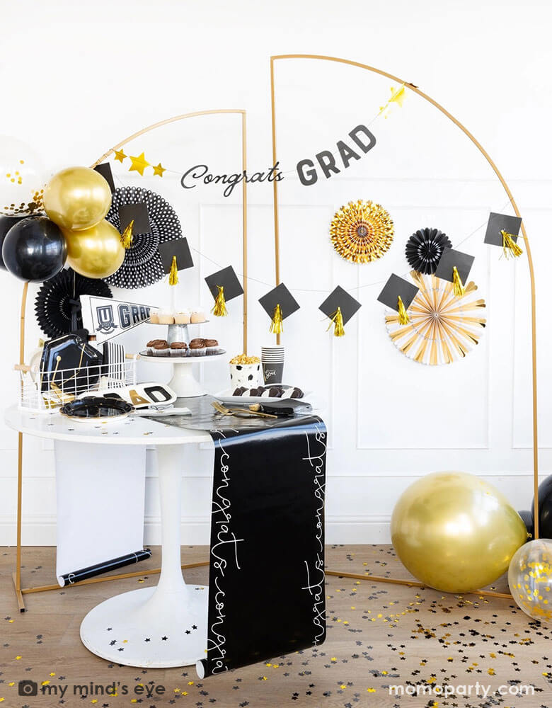 A graduation party featuring Momo Party's graduation party supplies by My Mind's Eye including U did it Grad felt party pennant, black striped dinner napkins, graduation cap shaped plates. On the table there are some cupcakes, cookies and popcorn on a black congratulation paper table runner and some black grad party cups and grad cap shaped napkins. In the back there are a gold backdrop frame adorned with some black and gold balloons and party fans, along with a Congrats Grad and grad cap tassels banner.