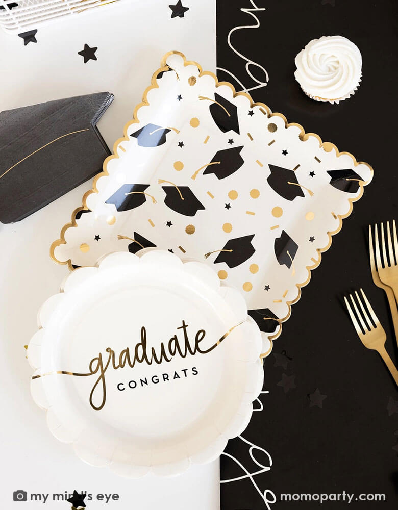 A graduation party table features Momo Party's 9" x 9" black and gold cap scatter paper plates, scalloped congrats graduate paper plates, grad cap shaped napkins and congratulation black paper runner along with some black and gold star shaped confetti by My Mind's Eye. Along with some cupcakes and gold utensils making this a great inspo for a modern and stylish graduation party tablescape.