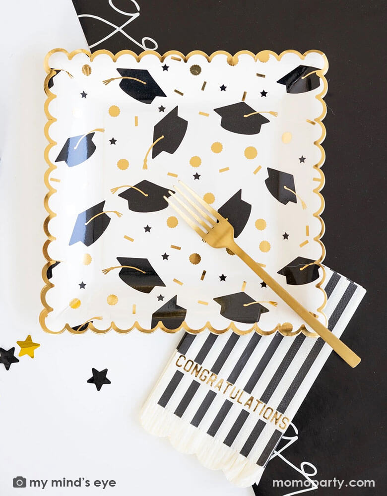 A graduation party table features Momo Party's 9" x 9" black and gold cap scatter paper plates, congratulations black and white striped dinner napkins, and congratulation black paper runner along with some black and gold star shaped confetti by My Mind's Eye. Making this a great inspo for a modern and stylish graduation party tablescape.