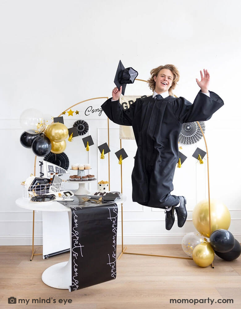 A graduate in his robe and cap jumping in a graduate party set up featuring Momo Party's graduation supplies including U did it Grad felt party pennant, black striped dinner napkins, graduation cap shaped plates. On the table there are some cupcakes, cookies and popcorn on a black congratulation paper table runner and some black grad party cups and grad cap shaped napkins. In the back there are a gold backdrop frame adorned with some black and gold balloons and party fans, along with a Congrats Grad banner.
