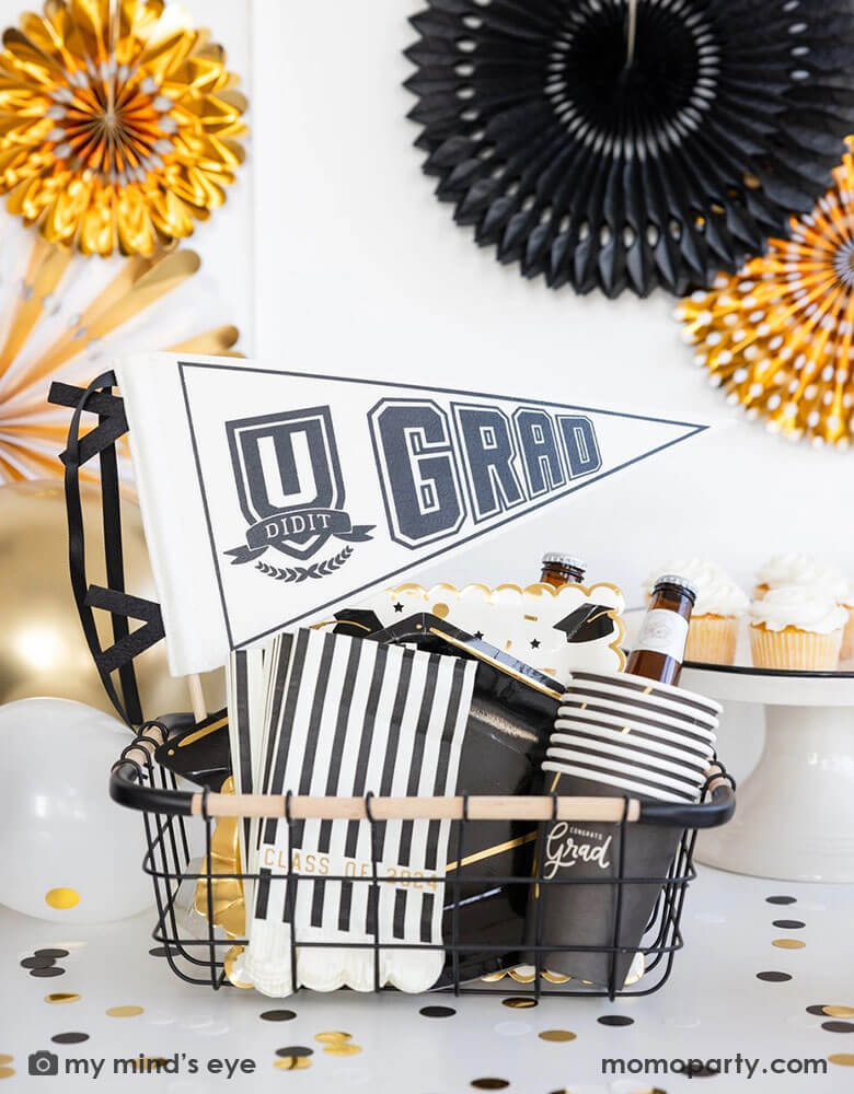 A graduation party set up featuring an occasion bin filled with Momo Party's graduation party supplies by My Mind's Eye including U did it Grad felt party pennant, class of 2024 black striped dinner napkins, graduation cap shaped plates, 12 oz grad party cups and grad cap scattered paper plates. In the back there are black and gold party fans hung on the wall and some cupcakes and graduation confetti on the table. Making this a perfect inspo for modern and stylish graduation party set up.