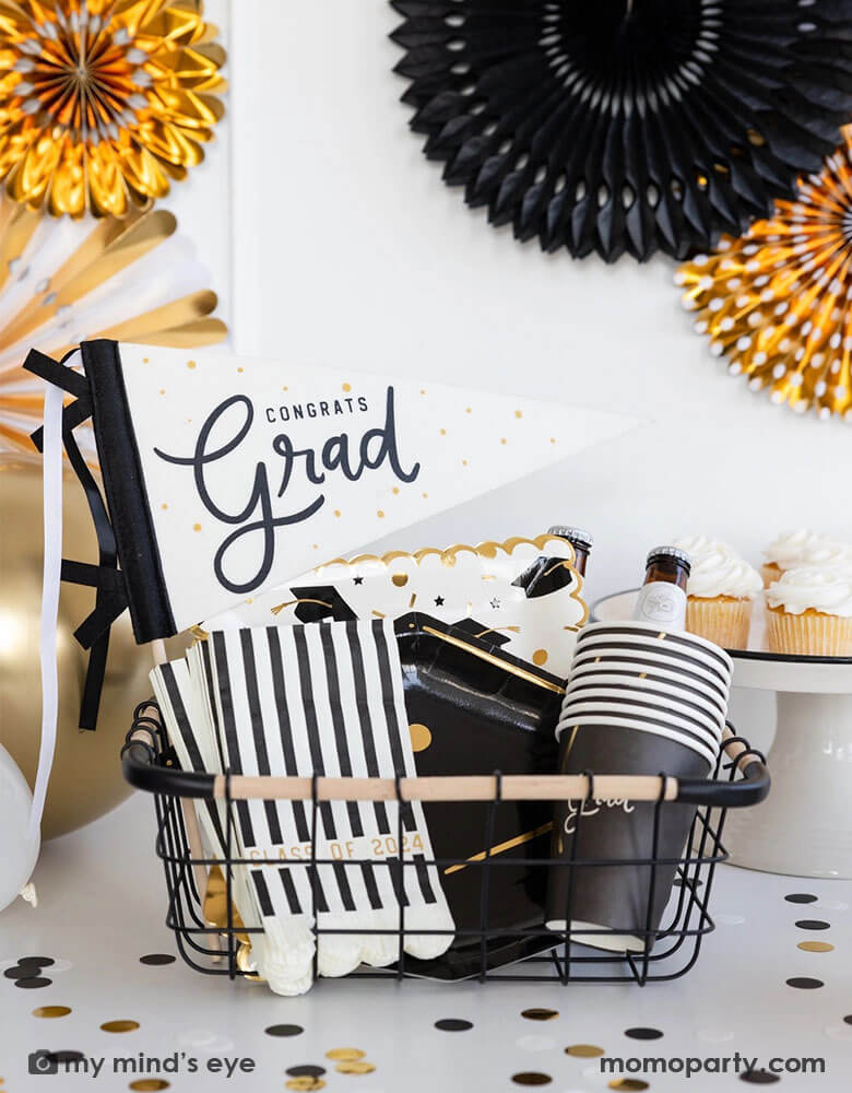 A graduation party set up featuring an occasion bin filled with Momo Party's graduation party supplies by My Mind's Eye including Congrats Grad felt party pennant, class of 2024 black striped dinner napkins, graduation cap shaped plates, 12 oz grad party cups and grad cap scattered paper plates. In the back there are black and gold party fans hung on the wall and some cupcakes and graduation confetti on the table. Making this a perfect inspo for modern and stylish graduation party set up.