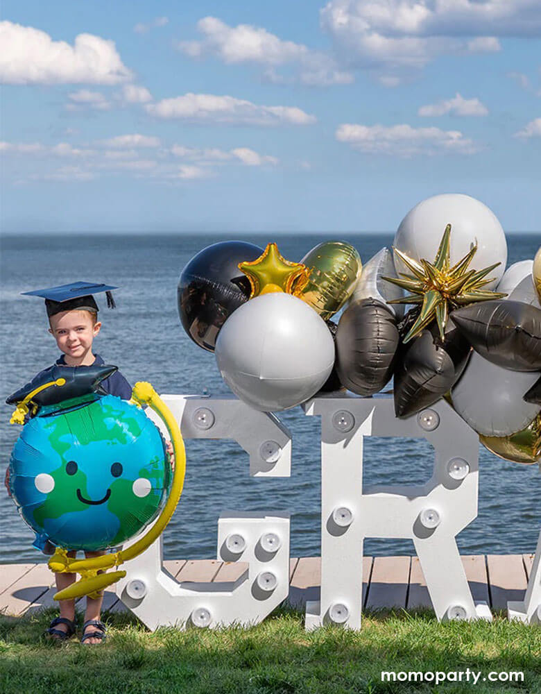 A preschool or kindergarten age kid holding Momo Party's 29" Graduation Fun Globe Shaped Foil Balloon standing next to a graduation marquee sign decorated with graduation themed balloon garlands in black, gold and white to celebrate his graduation day.
