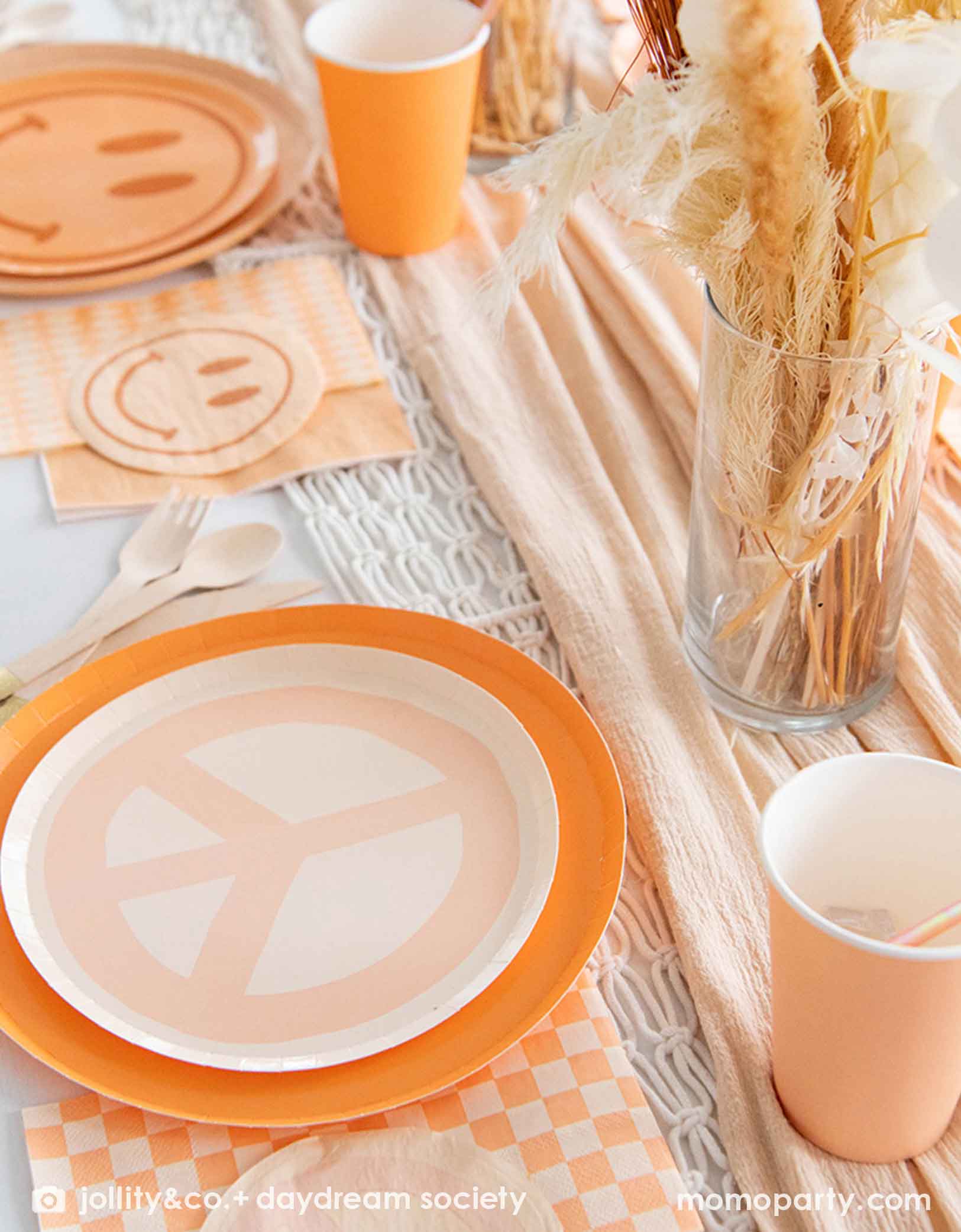 A neutral table setting with pastel colors of orange and peach peach sign and smiley face plates, peach cups, and checkered napkins in groovy hippie love and peace theme from Momo Party. There are also some dry leaves and feathers over a soft peach scarf as table centerpiece, making this a bohemian inspired tablescape.