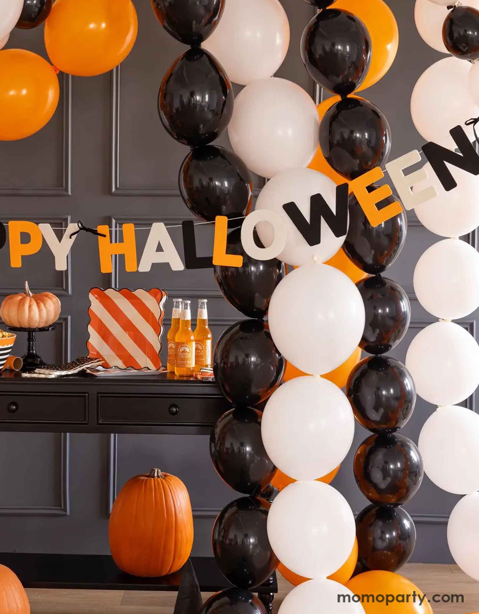A Halloween-themed party setup with balloons in the classic Halloween colors of black, white and orange, along with Momo Party's Happy Halloween felt banner decoration, and multiple Halloween themed tablewares including plates, napkins, serving bowls and more.