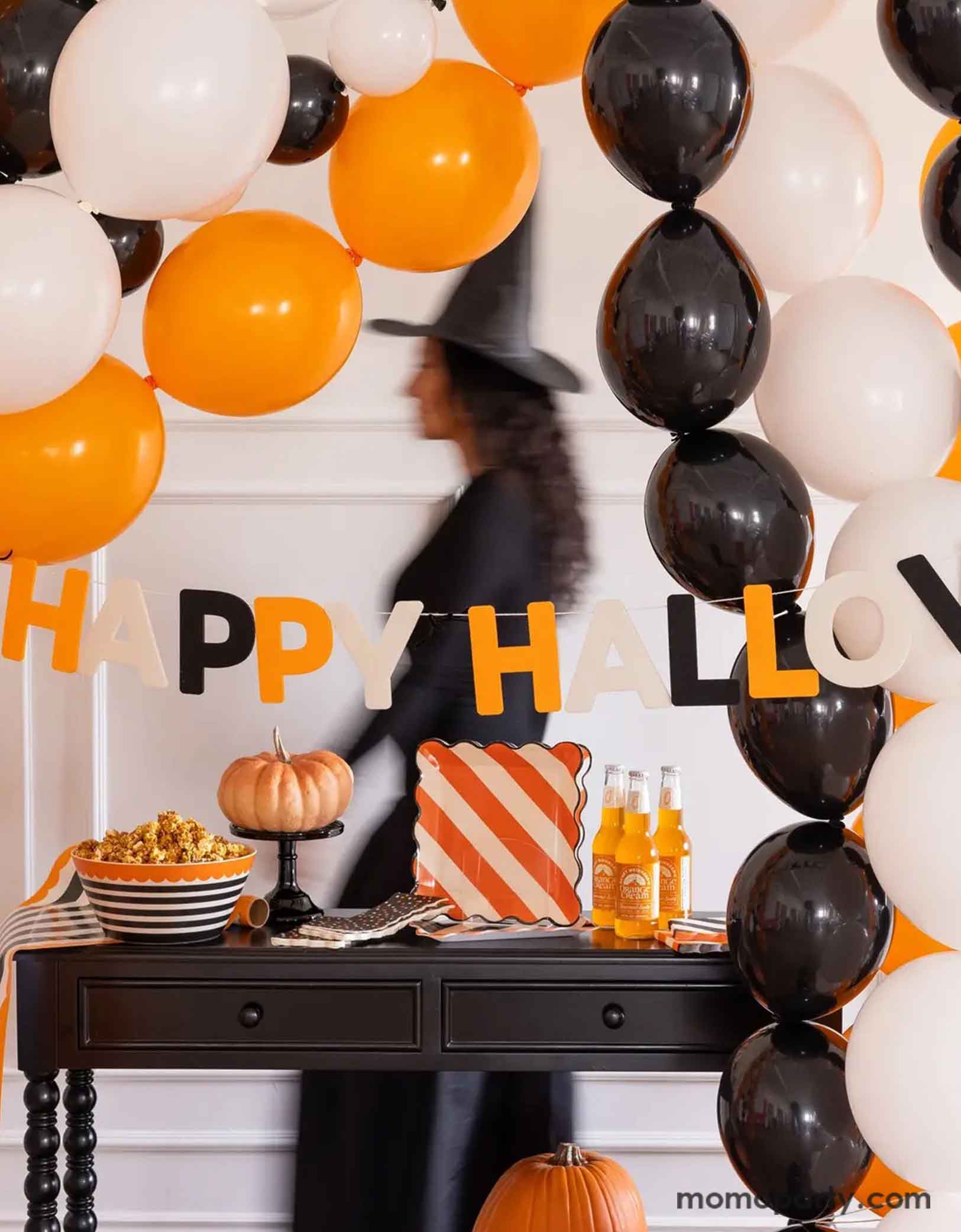 A Halloween-themed party setup with balloons in the classic Halloween colors of black, white and orange, along with Momo Party's Happy Halloween felt banner decoration, and multiple Halloween themed tablewares including plates, napkins, serving bowls and more. A lady in a witch costume walking by in motion in the back, making this a spooky yet fun inspo for a Halloween celebration.