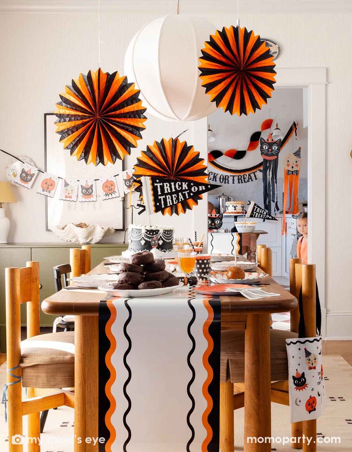 A styled Halloween dining room scene featuring various vintage Halloween themed party supplies and decorations from Momo Party with orange and black paper fan decorations hanging above a wooden table set with plates, drinks, treats and a white table runner with black and orange wavy stripes, along with a "trick or treat" party pennant in the middle. A doorway in the background shows more party decorations, including a vintage Halloween themed banner, bunting garland and hanging cat and ghost figures.
