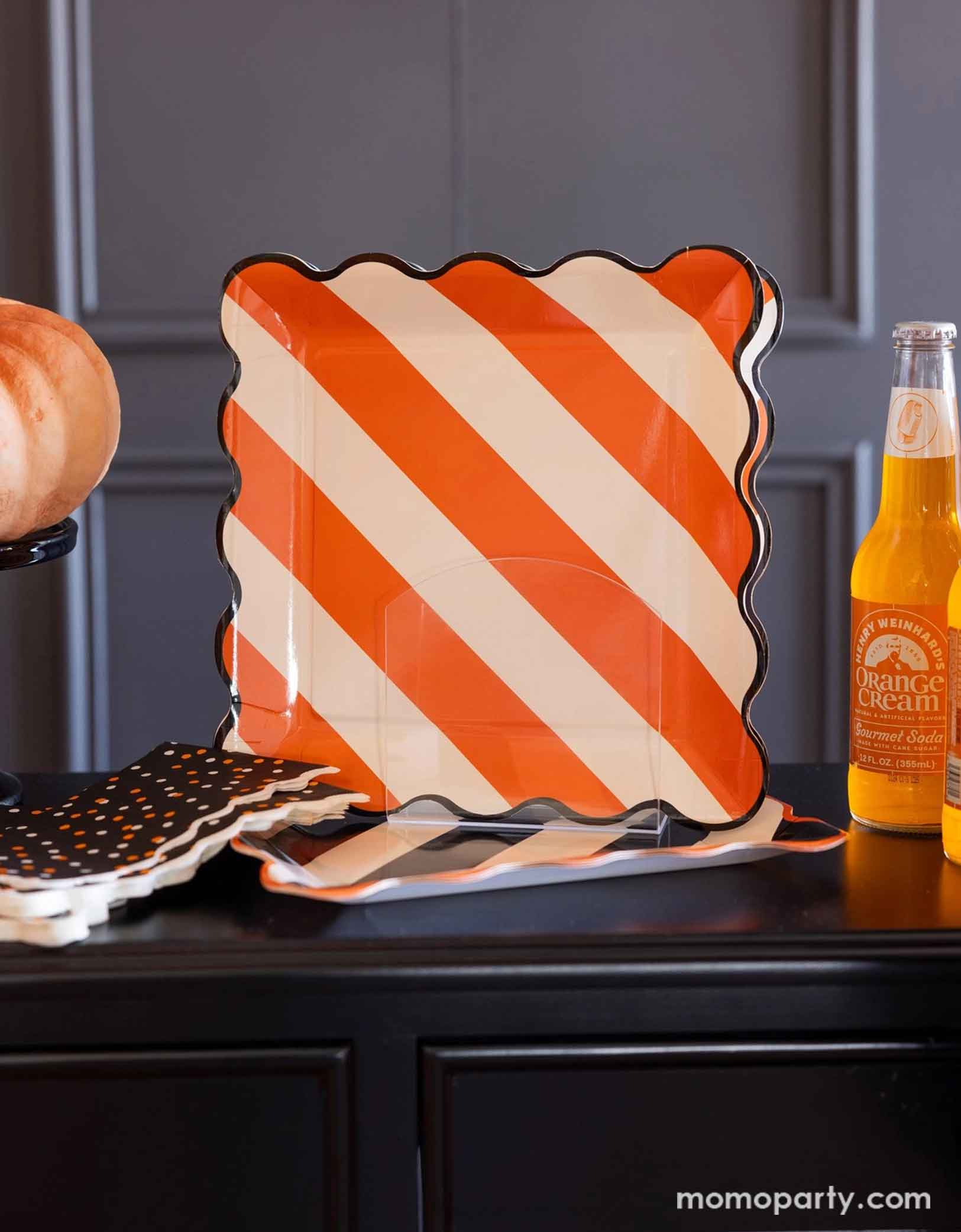 A black tabletop featuring Momo Party's Orange  Striped Plate with orange and white diagonal stripes with wavy black edge. Around the plate there are some Halloween-themed items including orange soda bottles, pumpkins, black star confetti napkins.