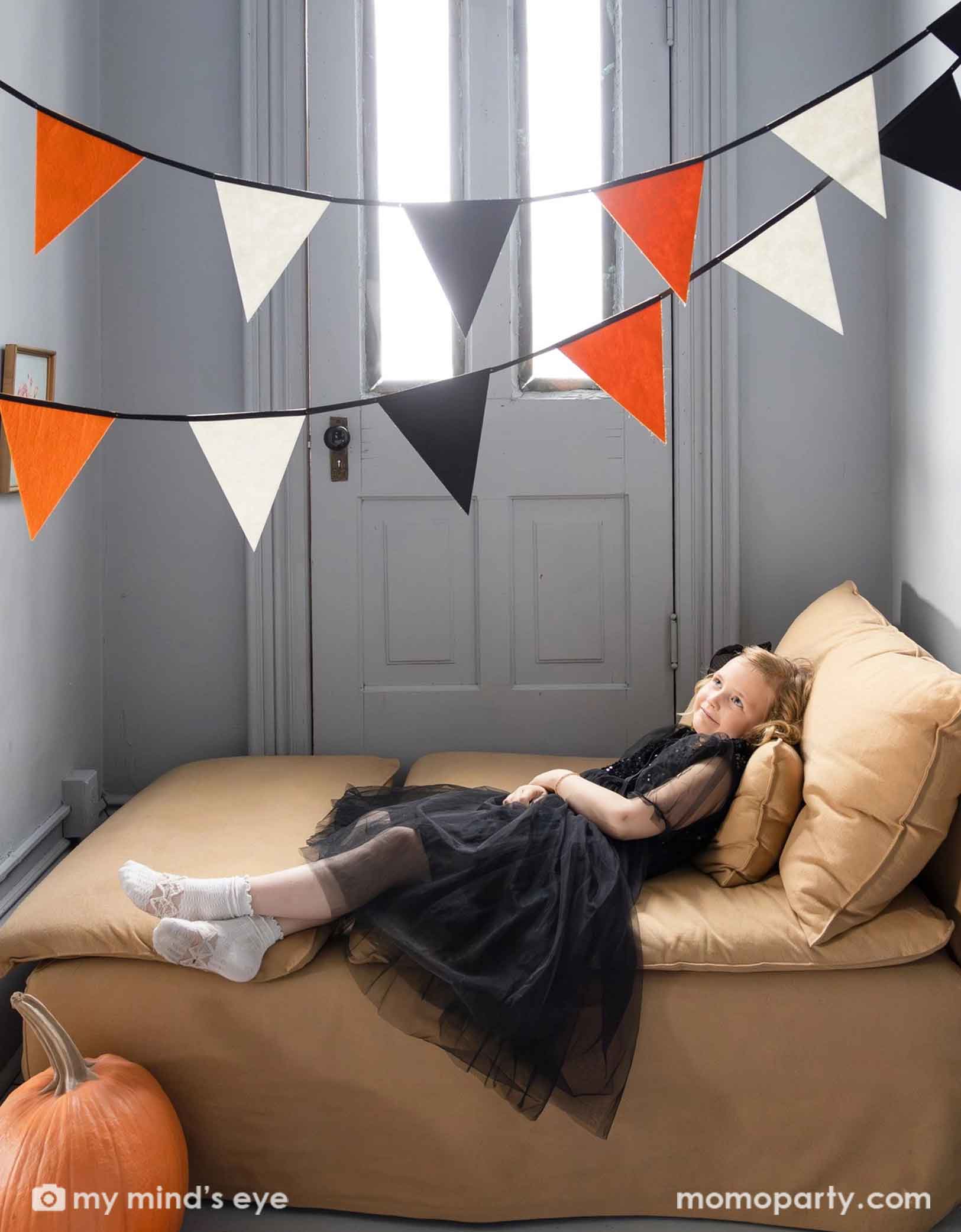 A girl in her Halloween black dress sitting on a beige couch next to a door with Halloween-themed decorations, including a pumpkin and  Momo Party's 6 ft orange, black and white flag banner hung on top.