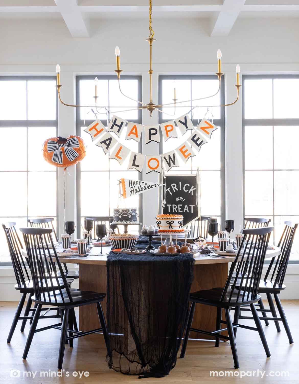 A styled Halloween dining room scene with a round wooden table covered in a black lace tablecloth, surrounded by black spindle-back chairs. A hanging banner reads “HAPPY HALLOWEEN,” with a cake on top, and a small sign that says “Trick or Treat.