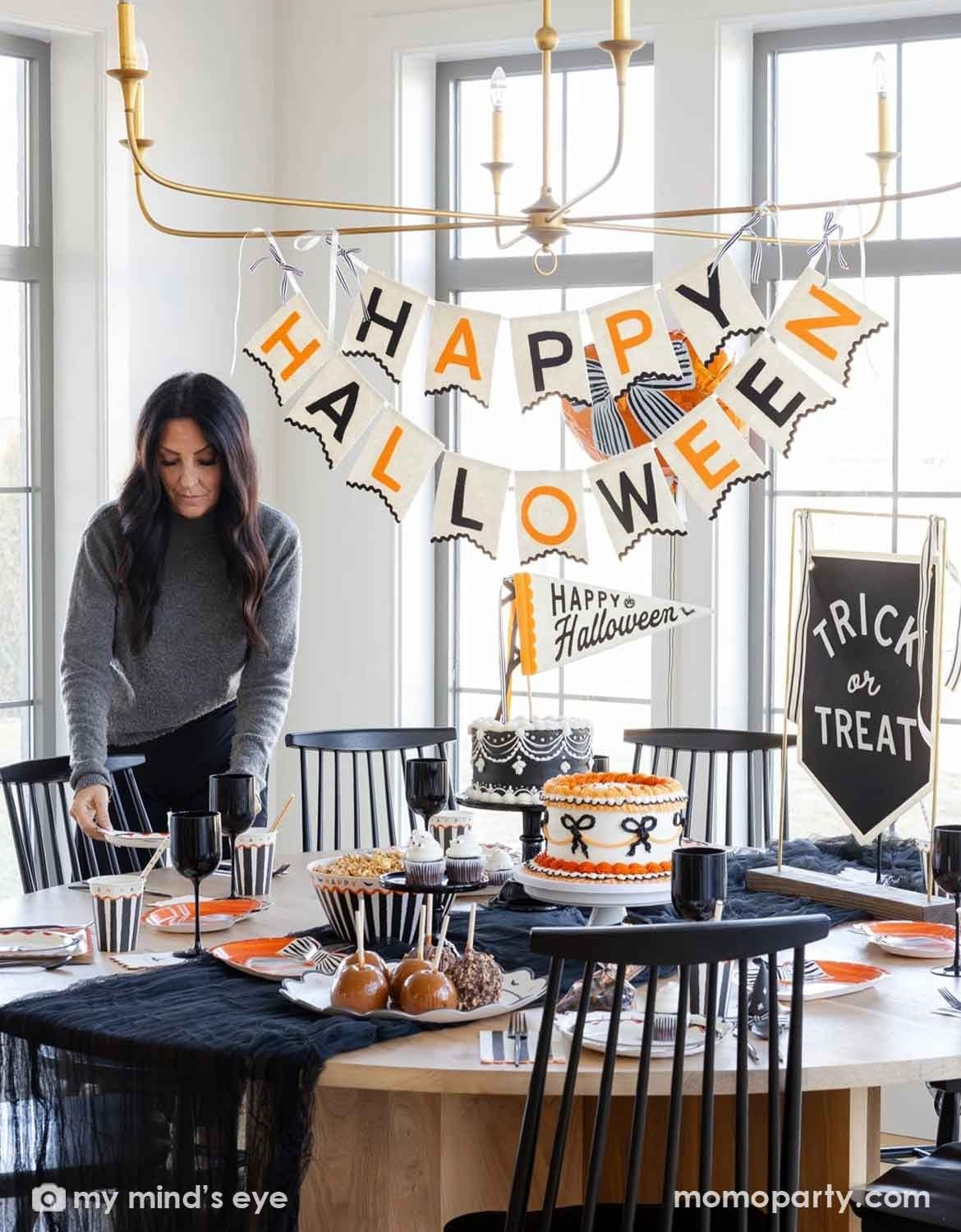A woman sets a Halloween table in a bright dining room. A styled Halloween party table setup in a bright dining room. Momo Party's Halloween large banner reading HAPPY HALLOWEEN hangs above the table, with a smaller sign that says Happy Halloween and a chalkboard sign that says Trick or Treat. On the table various Halloween themed party tablewares are featured, including party cups, plates, napkins and serving bowls - making this a modern and elegant Halloween party decorating inspo.