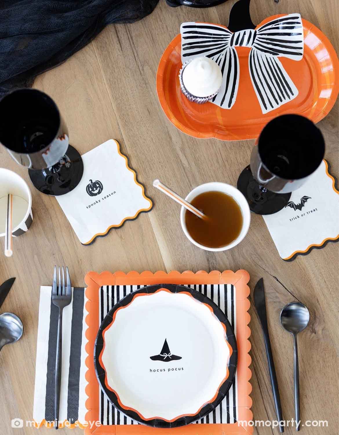 A styled Halloween table setting on a light wood table, featuring orange and black scalloped place settings, black goblets, white napkins with black jack-o'-lantern and bat motifs, and Momo Party's pumpkin shaped plate with a black and white bow on it.