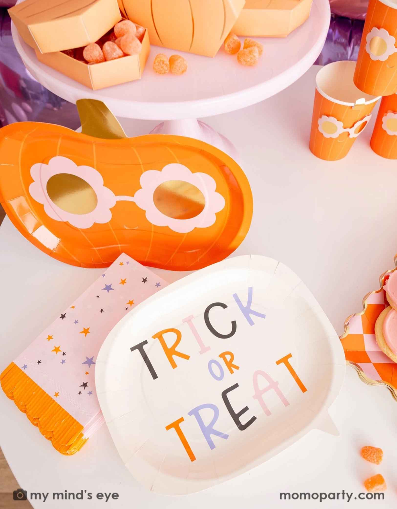 A Halloween-themed table featuring various pink Halloween themed tableware from Momo Party including a 'Trick or Treat' plate, the groovy pumpkin-shaped plate and party cups, plus pink star with orange fringed napkins. Making this an adorable tablescape for a cute Halloween party celebration.