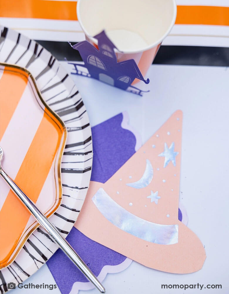 A close up shot of a festive Halloween witch themed party table featuring Momo Party's Bewitching orange side plate on a black and white striped dinner plates, bewitching witch shaped hat napkin and a party cup decorated with haunted house, on a black and orange striped table runner. A perfect inspo for Halloween witch themed table for this spooky season!
