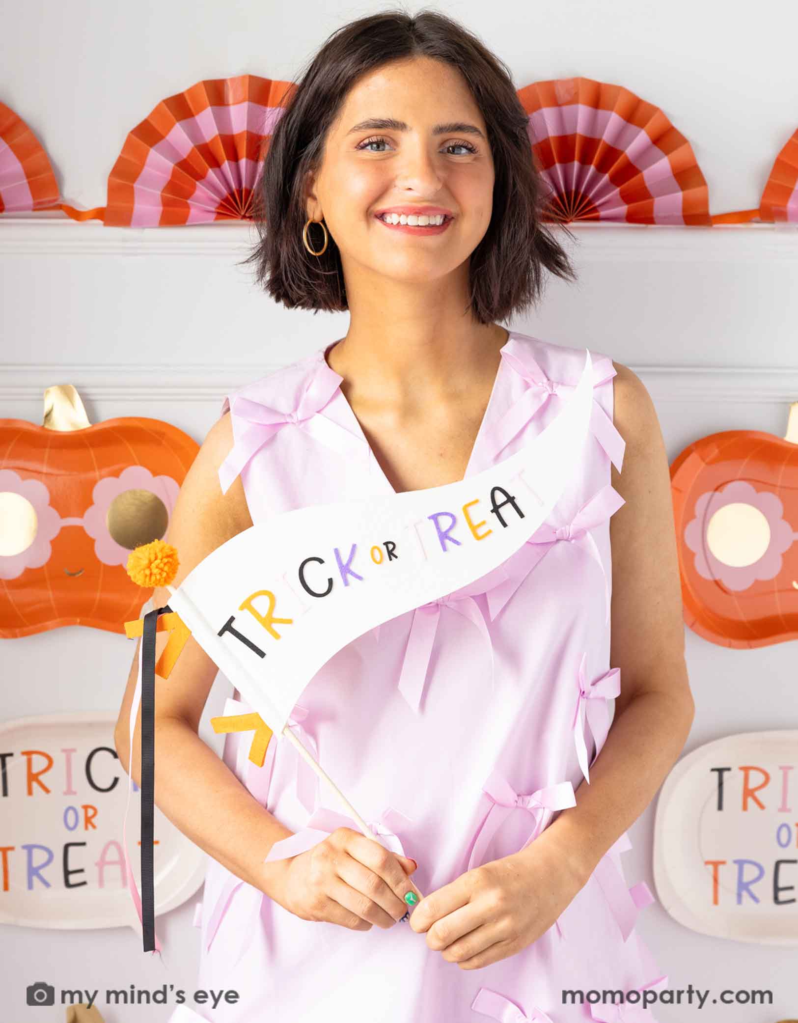 A lady in a pink dress holding Momo Party's 14" 'Trick or Treat' felt party pennant smiling into the camera with Halloween decorations on the wall in the background.