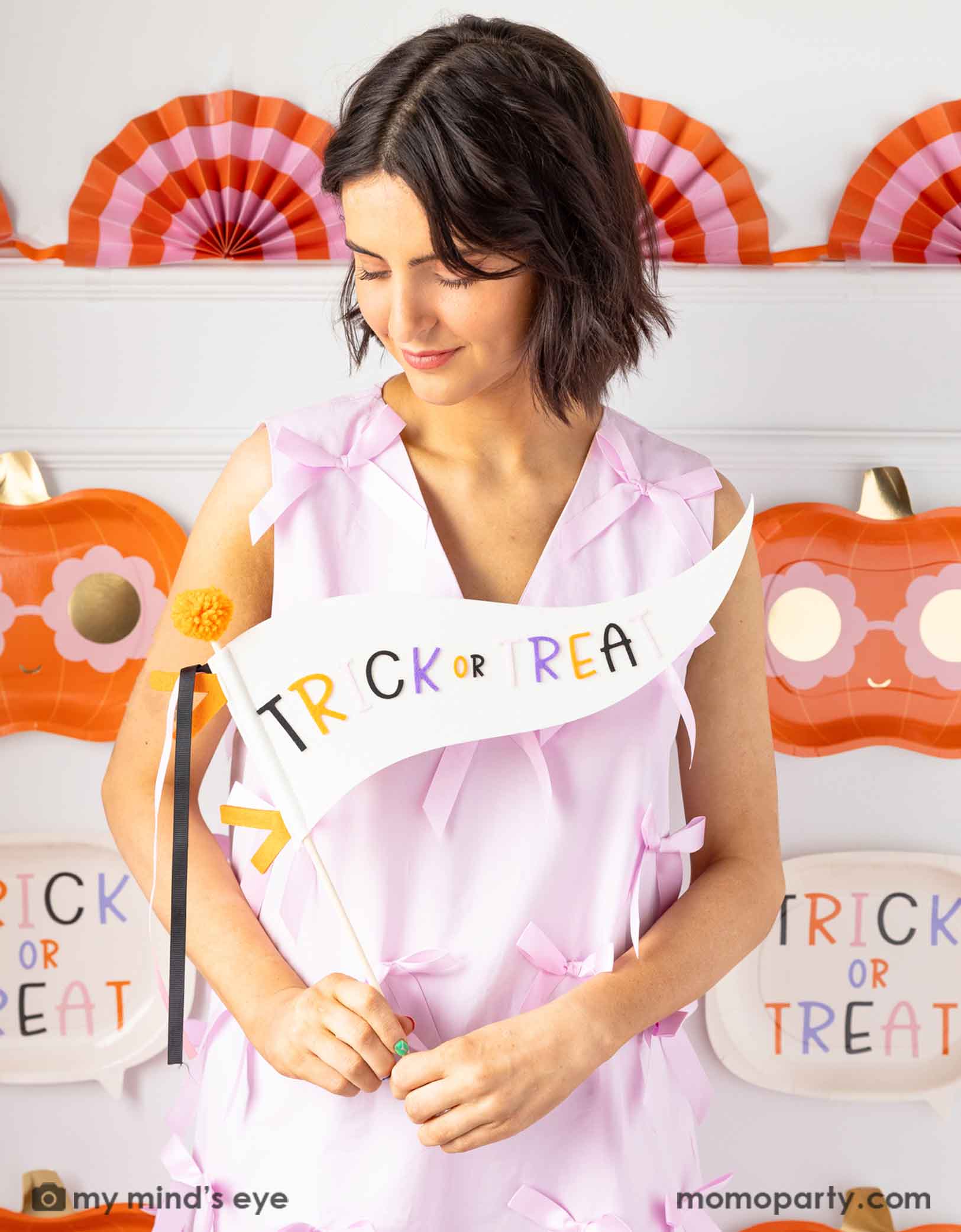 A lady in a pink dress holding Momo Party's 14" 'Trick or Treat' felt party pennant with Halloween decorations on the wall in the background.