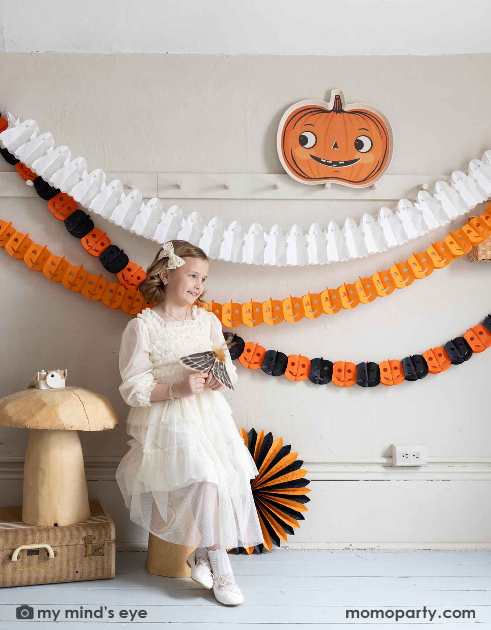 A girl in a vintage inspired white tulle dress sitting next to a white wall with coat hanging rack decorated with Halloween-themed decorations including Momo Party's pumpkin-shaped bamboo tray, ghost tissue garland, orange Jack-o'-lantern tissue garland, plus an orange and black Jack-o'-lantern tissue garland hung on the wall. Underneath is a cat figurine on a bunch of wooden mushroom stools which are next to a orange and black tissue paper fan decoration. 