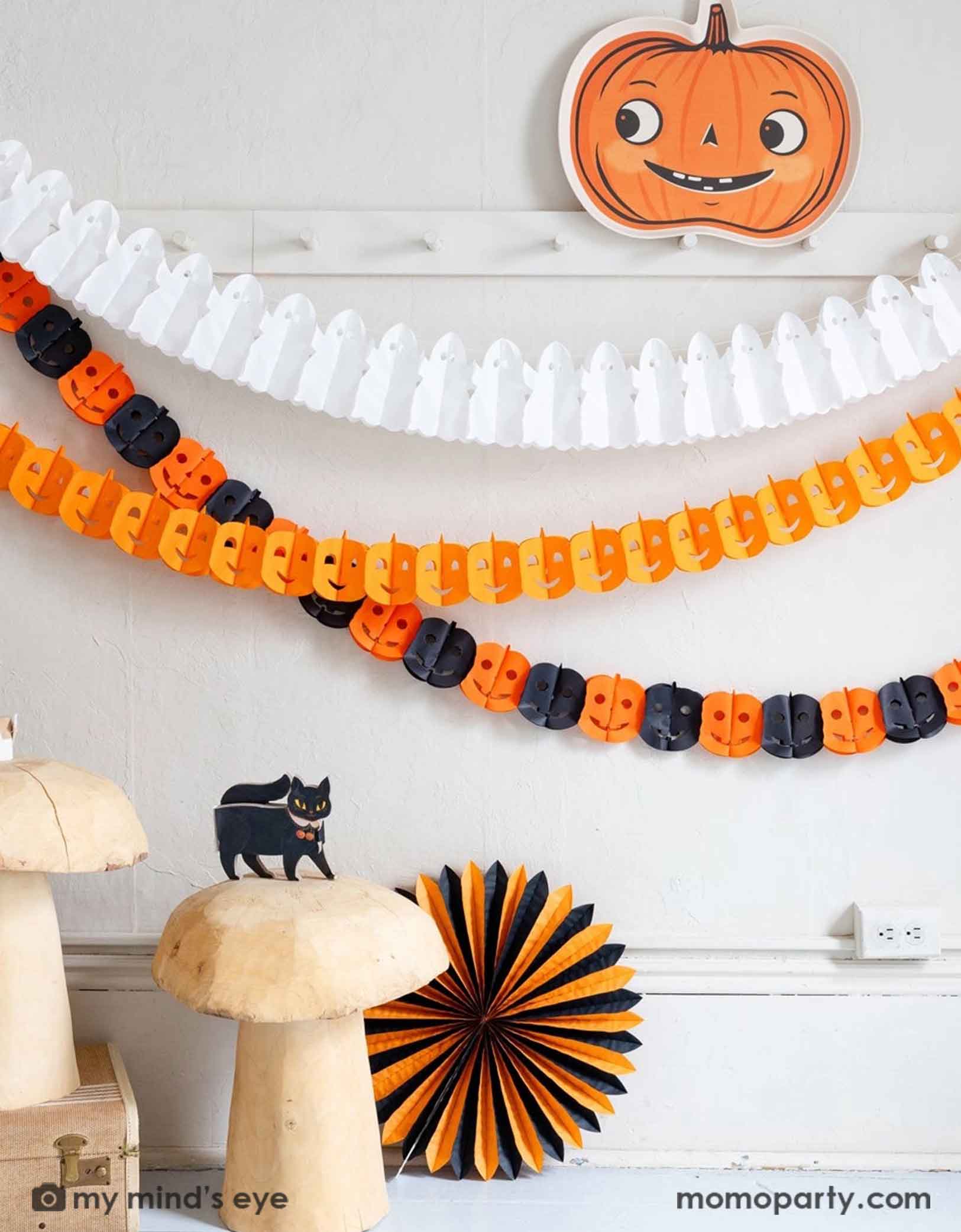 A white wall with coat hanging rack decorated with Halloween-themed decorations including Momo Party's pumpkin-shaped bamboo tray, ghost tissue garland, orange Jack-o'-lantern tissue garland, plus an orange and black Jack-o'-lantern tissue garland hung on the wall. Underneath is a cat figurine on a bunch of wooden mushroom stools which are next to a orange and black tissue paper fan decoration. A simple and modern way to decorate your home in the Halloween spooky season.