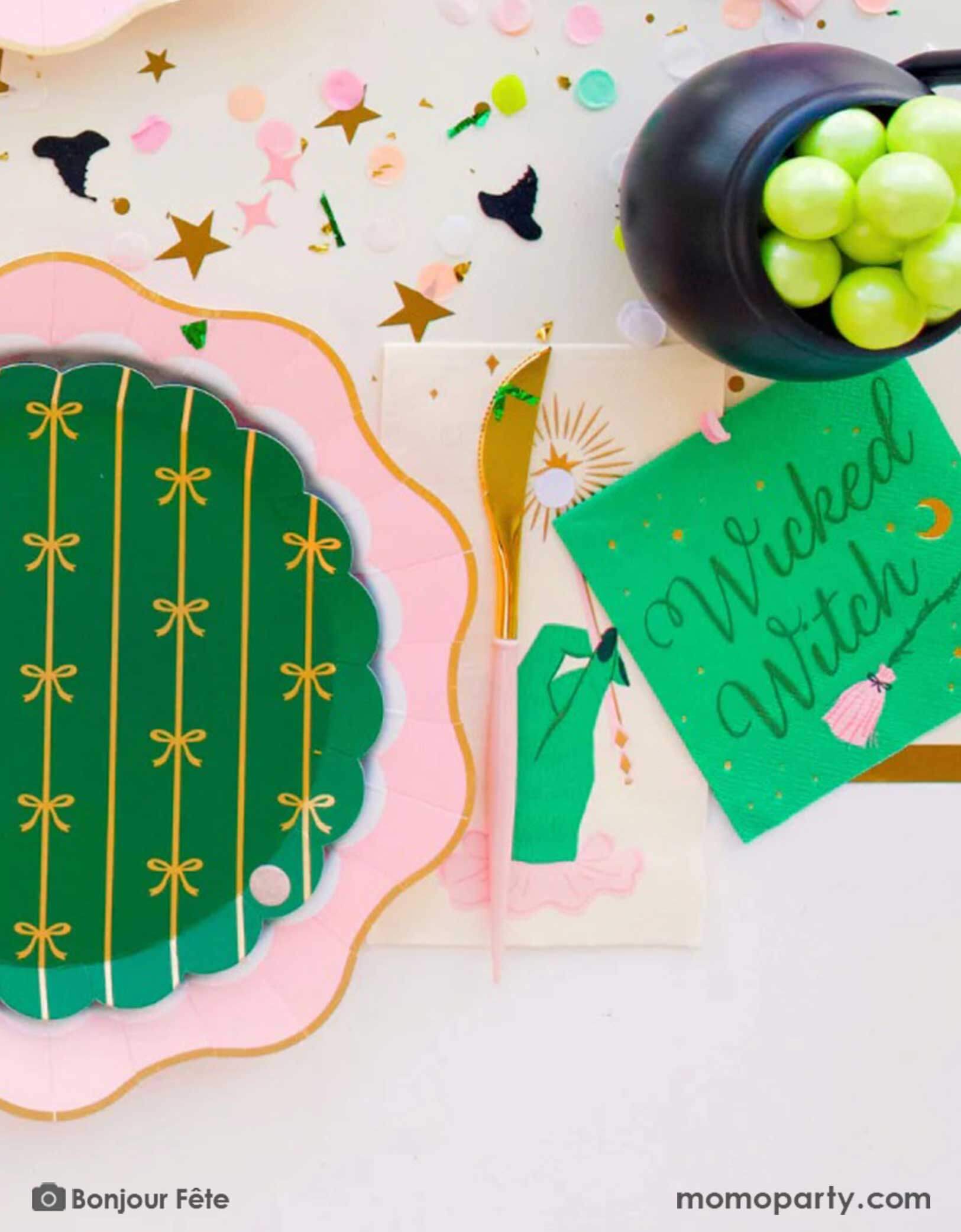 A whimsical Wicked themed party table setting featuring multiple Wicked themed party supplies from Momo Party including the Wicked Witch 5" small napkins, the magic wand guest napkins, a large pink dinner plate and an emerald green side plates with gold foil bows pattern, along with Wicked themed confetti spread out on the table and a mini cauldron filled with green grapes, it's a perfect party inspo for a fun and magical Wicked themed party in the Halloween spooky season.