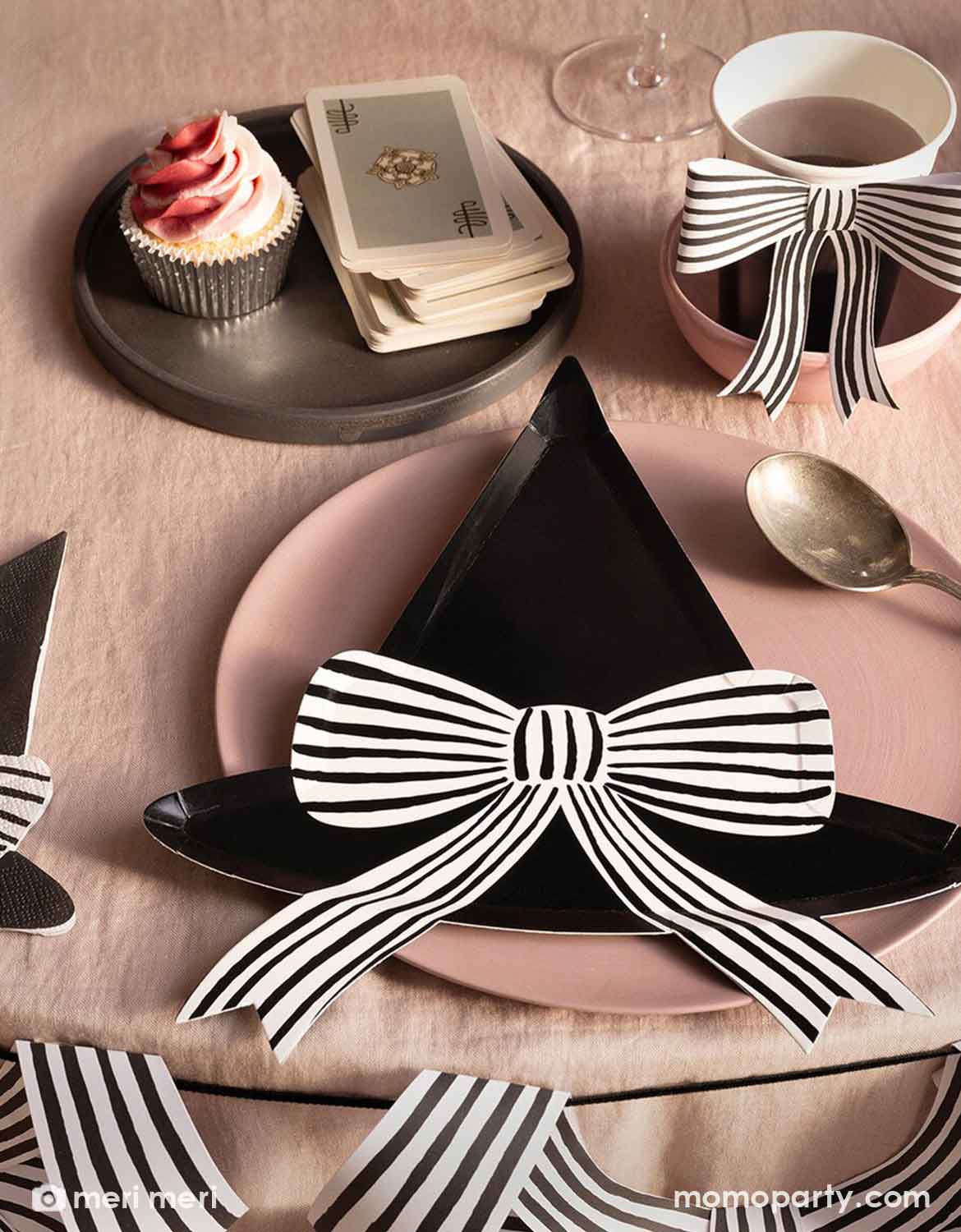 A styled pink Halloween party table setting featuring Momo Party's black witch hat shaped plate with a black and white striped bow on a pink plate, alongside a cup of tea with a matching striped bow, a cupcake, cards, and a spoon on a beige tablecloth adorned with black striped bow banner.