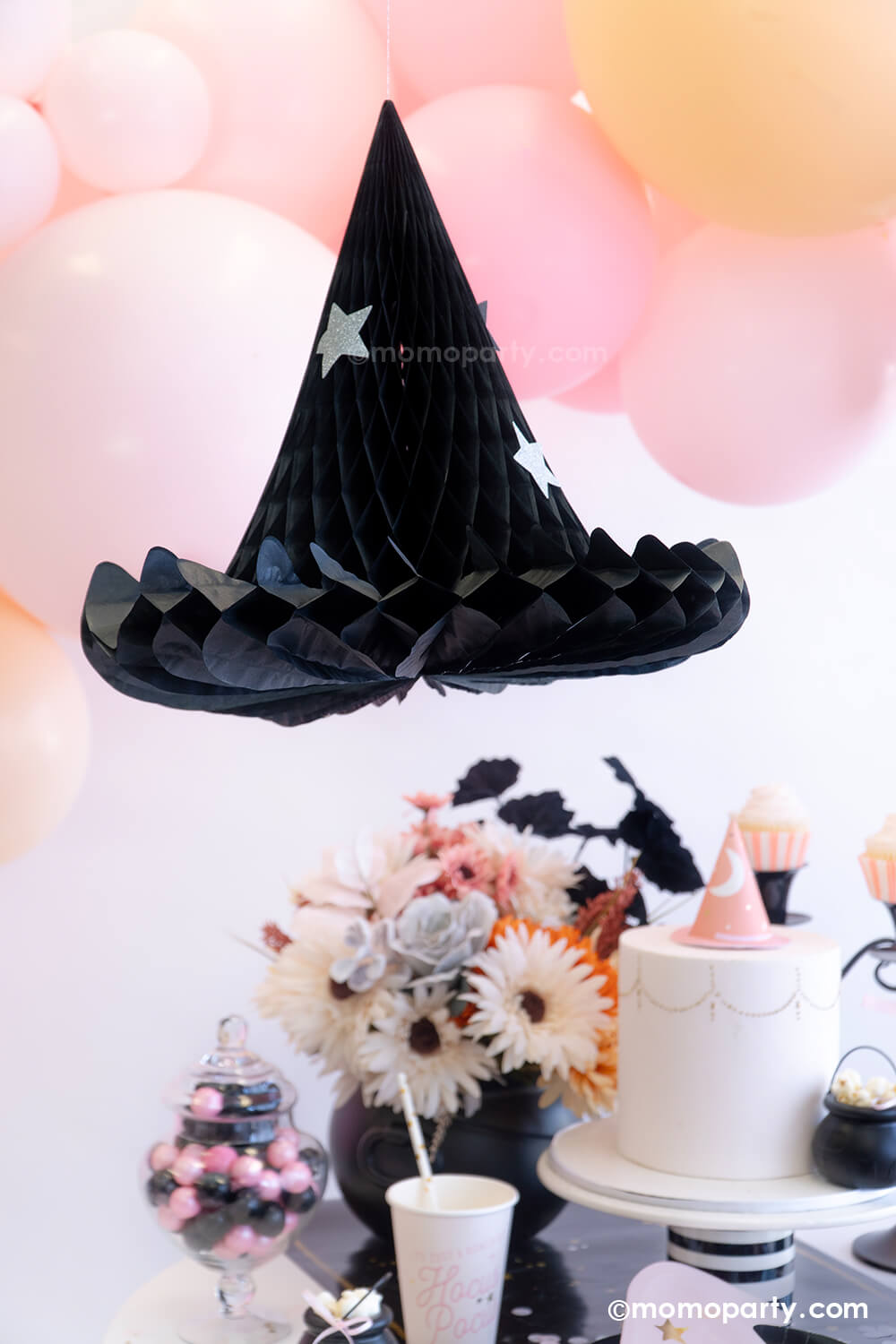 Momo Party's Hocus Pocus Witch Hat Honeycomb decoration hung next to an adorable pink balloon garland with various sized balloons in pastel pink, pink and blush colors. Below it is a pink inspired Halloween party table featuring witch themed party supplies from Momo Party including Hocus Pocus party cups, napkins, witch hat shaped plates and some pink and black themed treats. This makes a great inspo for a sweet girly pink Halloween that's perfect for this spooky season.