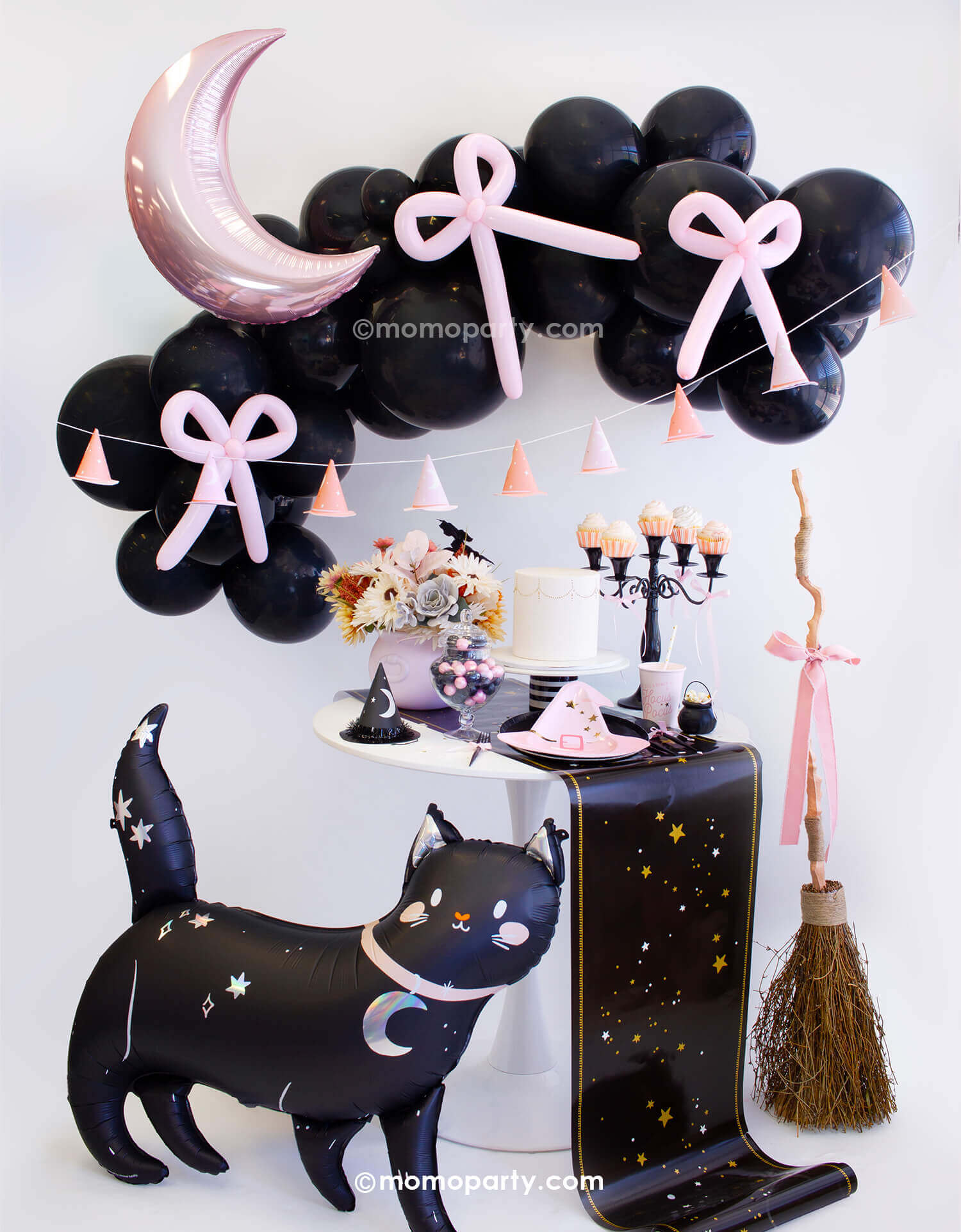 An adorable kid's friendly witch themed Halloween party look styled by Momo Party. With black balloon garland adorned with pastel pink balloon bows and the pink crescent moon shaped foil balloon, along with pink witch hat party banner as the backdrop. On the party table there are witch themed tableware including pink witch hat shaped plates, Hocus Pocus napkins and party cups, along with mini cauldrons, witch hats and treats. In the front there are a cute black cat foil balloon and a broom with a pink bow.