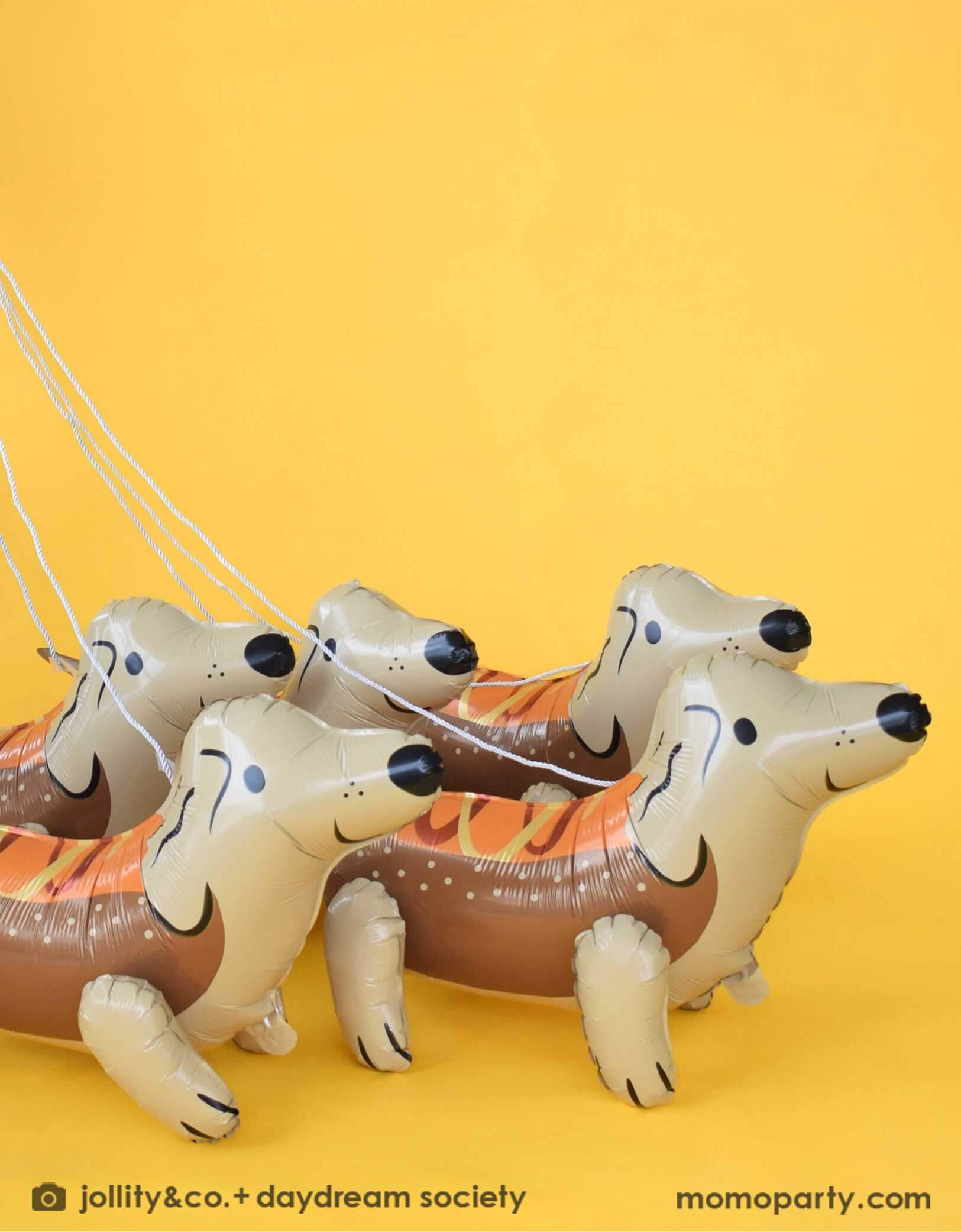 Four Momo Party's 26.8" x 16.5" x 12.6" Dachshund Dog-shaped foil balloons with a hot dog costume by Daydream Society on leashes on a yellow background.