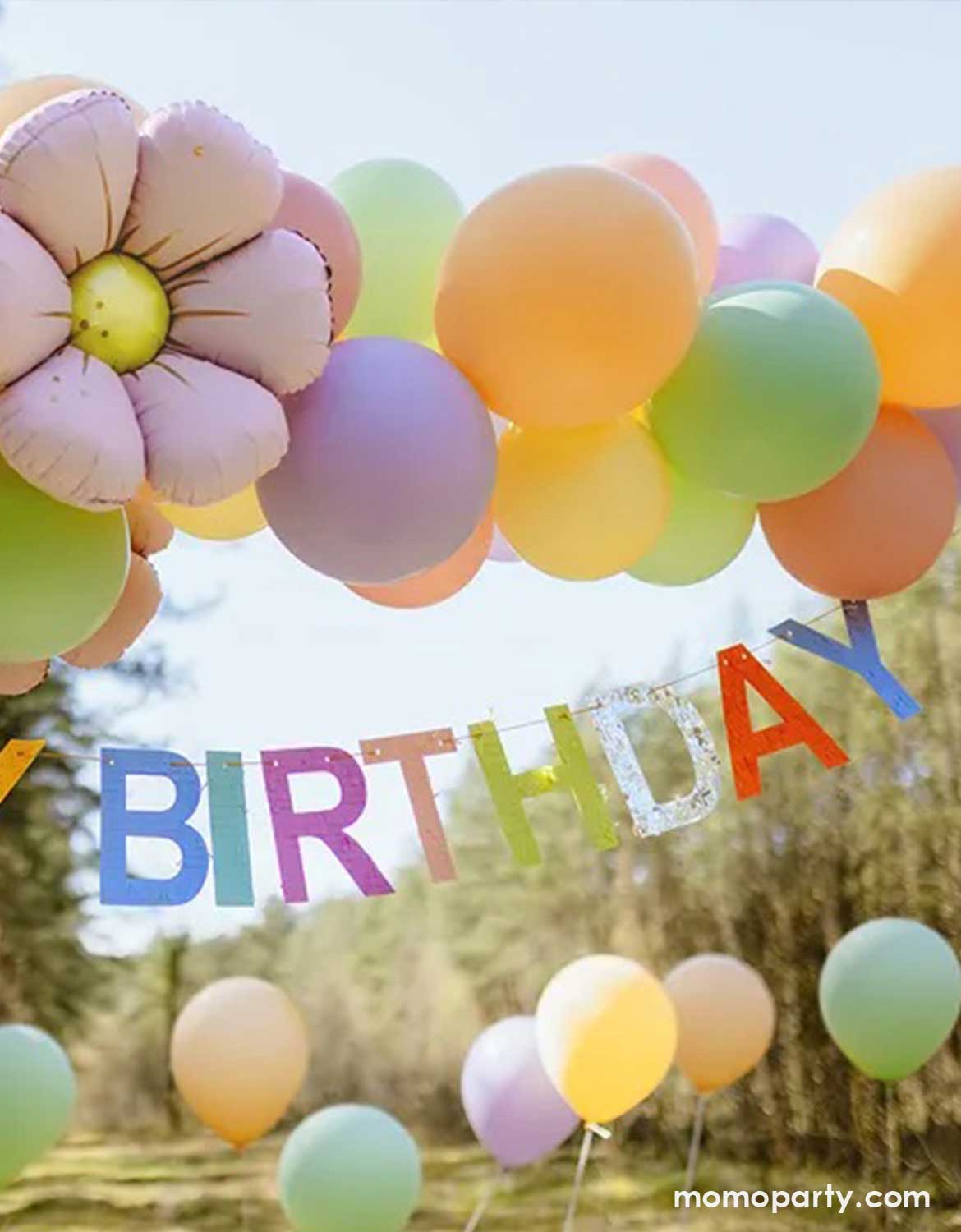 Colorful balloon arch in pastel colors with a lilac daisy-shaped foil balloon and Momo Party's  7ft 'Happy Birthday' banner in festive colors against a forest background.