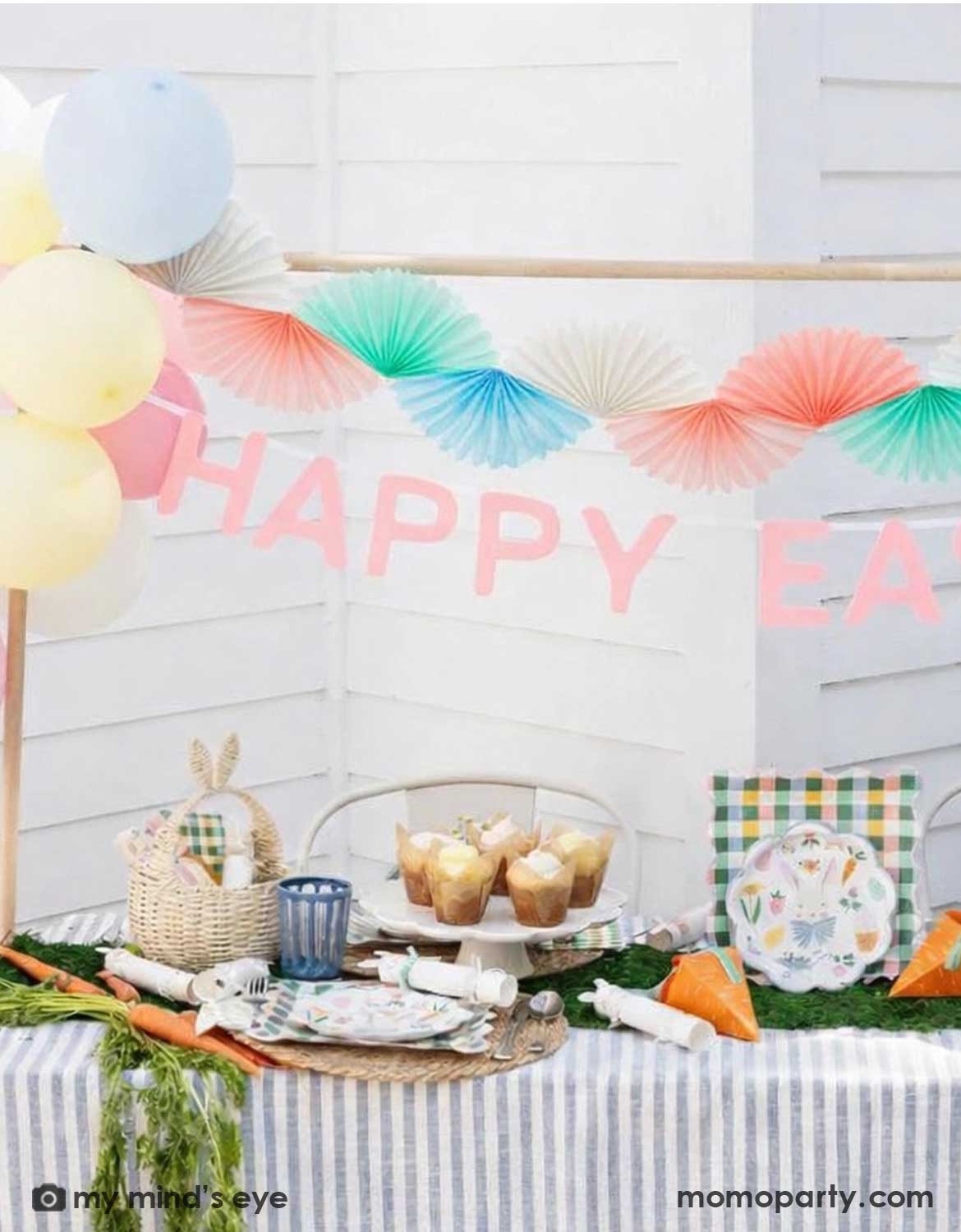 An Easter-themed table setting with multiple Easter themed tablewares on the table with colorful balloons, 'Happy Easter' banner, and Momo Party's pastel tissue bunting banner decorations hung above.