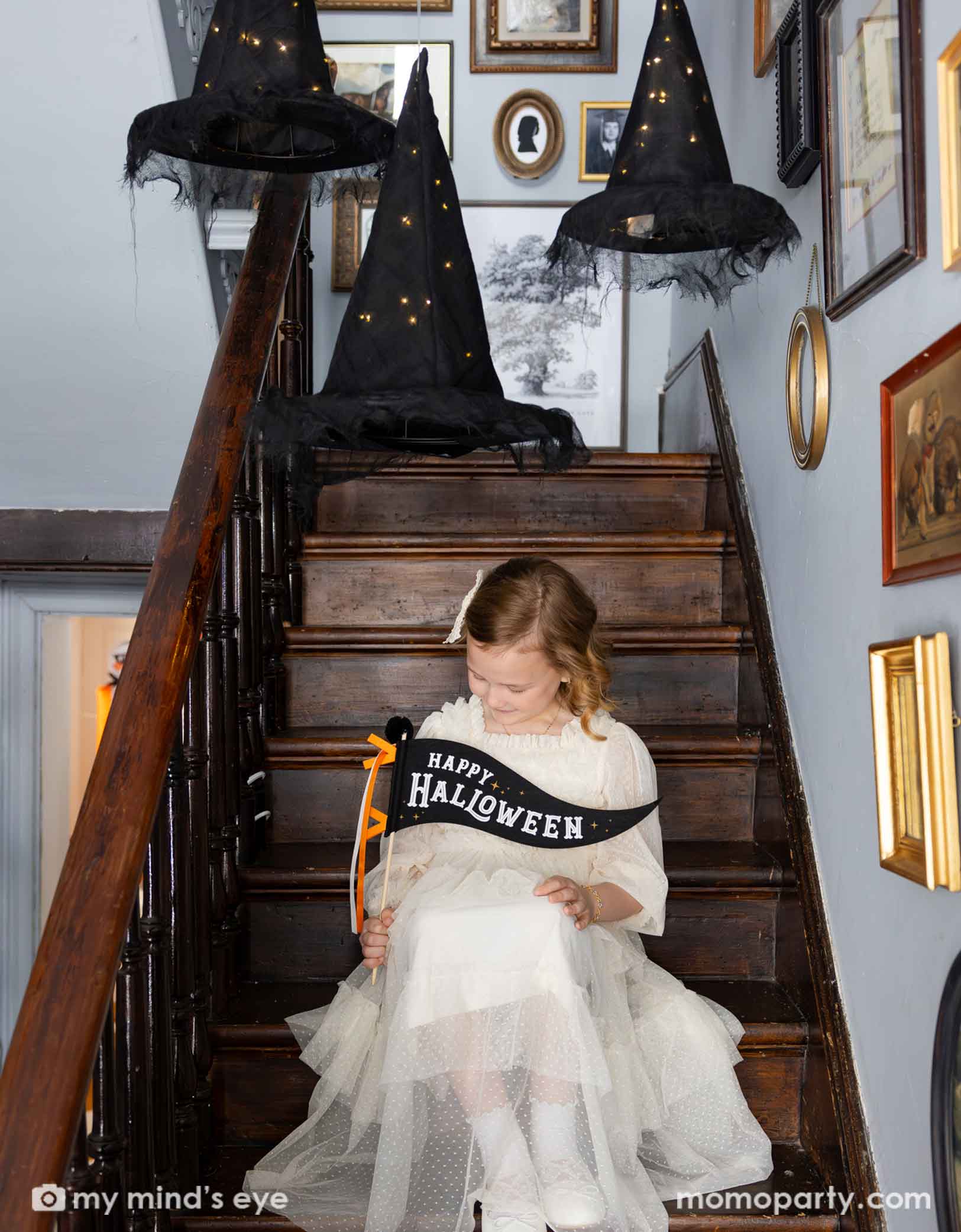 A girl in a vintage Halloween inspired white tulle dress sitting on a staircase holding Momo Party's 14" Happy Halloween felt party pennant looking down. Above her are three lit-up witch's hats hung from the ceiling. On the walls there are lots a vintage photo frame with old pictures, giving it a spooky vibe to a Halloween celebration.