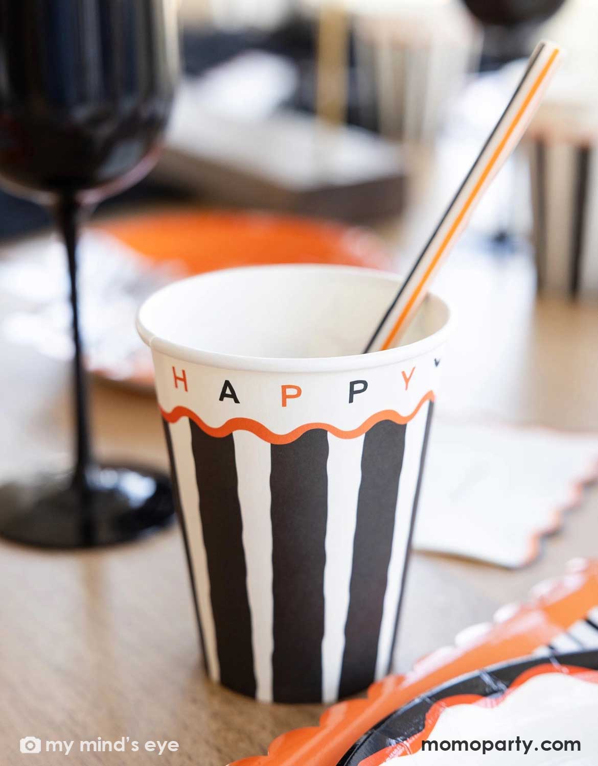 Momo Party's 12oz Halloween-themed paper cup with black and white vertical stripes, an orange wavy line, and the word 'HAPPY Halloween' printed on the side, shown on a Halloween party table with a striped straw. Around the cup there are various Halloween party tablewares including plates and napkins in orange and black colors. 