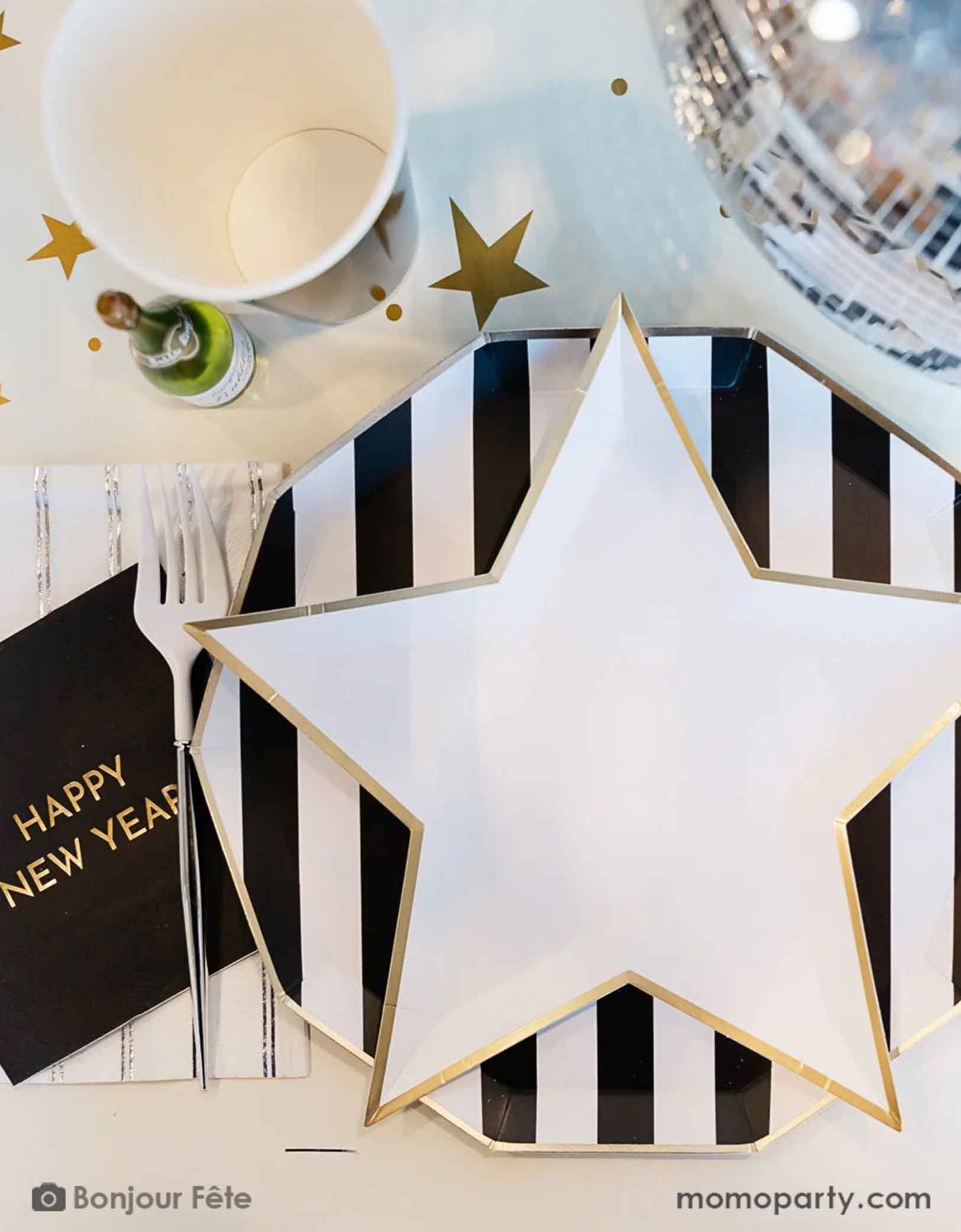 A white star-shaped plate with black and white striped plate and gold trim from Momo Party on a table setting with a black 'Happy New Year' napkin silver striped napkin and a mini champagne bottle on a gold star table rummer for a new year's party celebration.