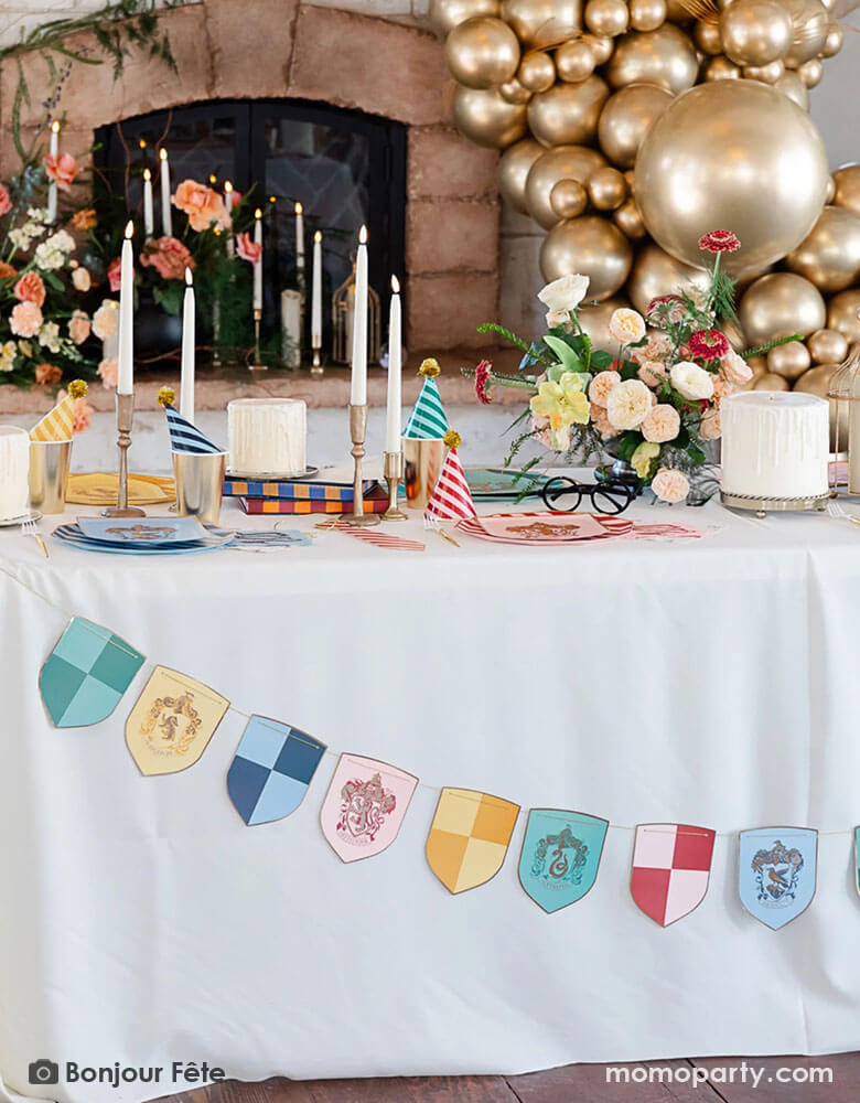 A Harry Potter themed party table adorned with Momo Party's Harry Potter school pride multicolor party banner featuring different Harry Potter house symbols and colors including Hufflepuff, Slytherin, Gryffindor and Ravenclaw. On the party table there are Harry Potter themed party tableware including plates, napkins, party hats, gold cups and white and gold candle sticks. In the back of the party table is a old vintage fireplace adorned with flowers and an organic gold balloon garland inspired by Quidditch.