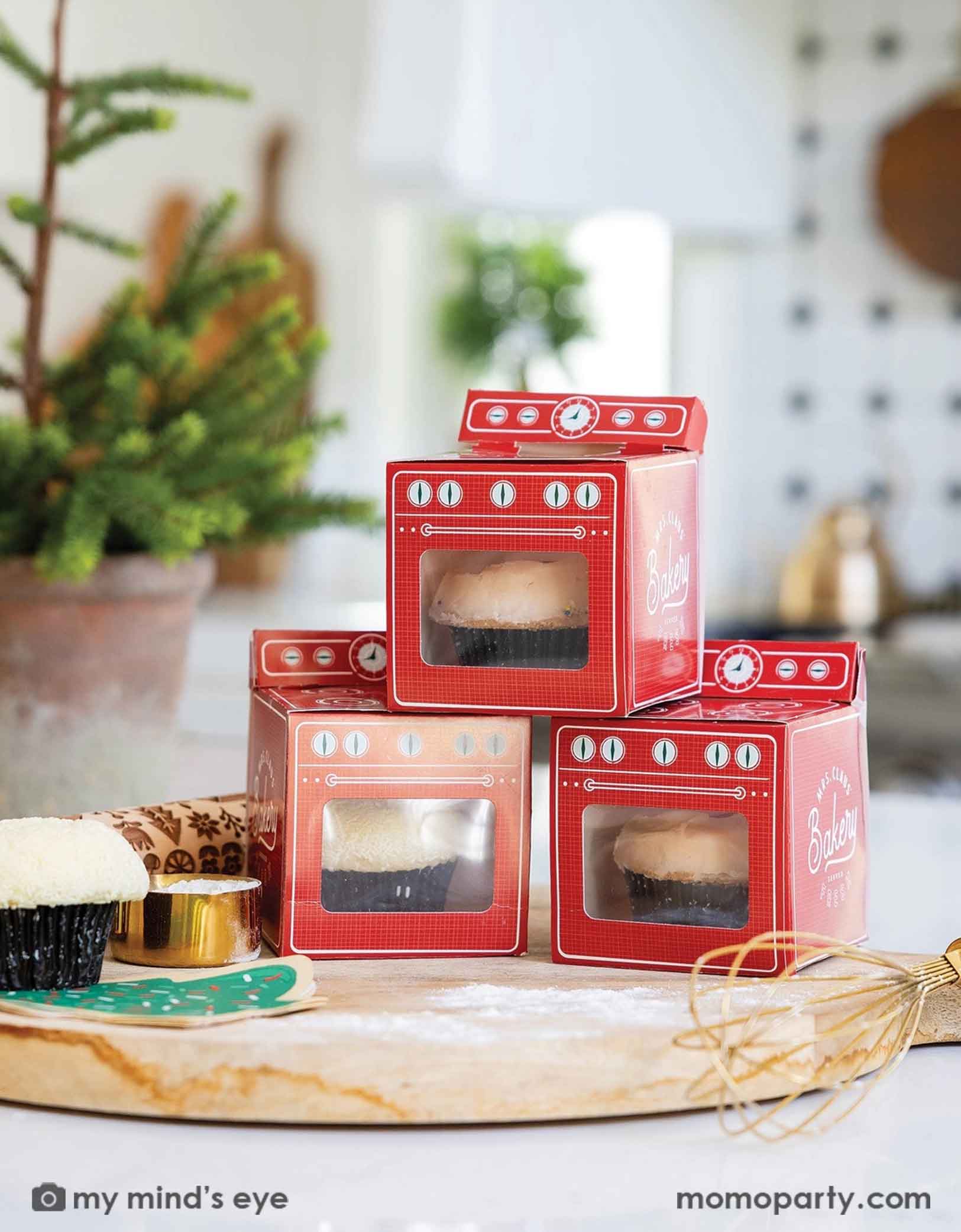 A Holiday Christmas themed decorated kitchen counter with Momo Party's 3.5" x 4" x 3.5" red vintage-style oven shaped treat boxes with cupcakes in them. Around the treat boxes are some baking utensils including a cutting board, a roller pin, a whisk with some flour around - a cute Holiday baking activity set up with kids!