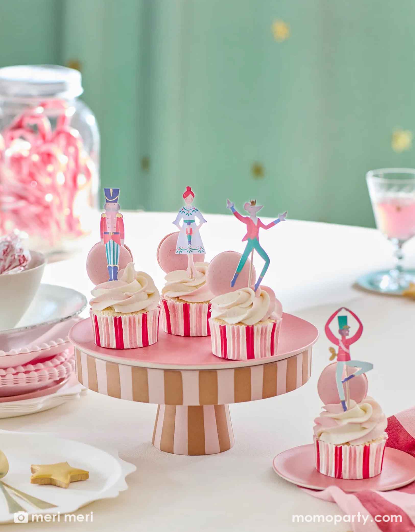A closeup shot of some cupcakes decorated with Momo Party's Nutcracker ballet cupcake kit featuring the Nutcracker  toppers on a striped cupcake liners on a striped cupcake stand with some pink plates, cookies, treats and candy canes in a candy jar on the table. In the back it's a green curtain adorned with little gold star stickers on it. Making this a festive and magical scene for the Holiday season.