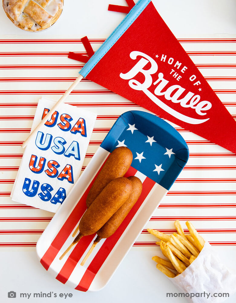 A festive patriotic themed party table featuring Momo Party's 15" Home of the Brave red felt pennant by My Mind's Eye, American flag shaped mini bamboo serving tray with summer snacks like corndogs and fries, next to it is a guest napkin with retro type of USA on it. With a matching red striped table runner, this makes a great inspo for a Memorial Day celebration or 4th of July Independence Day party.