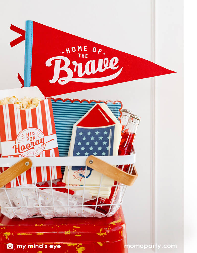 An occasion bin filled with festive patriotic themed party goods including Momo Party's 15" Home of the Brave red felt pennant by My Mind's Eye, blue stripe scallop-edged plates, rocket shaped guest napkins, red striped popcorn treat boxes with "hip hip hooray" on it, these are perfect decorations for a Memorial Day celebration or 4th of July Independence Day party!