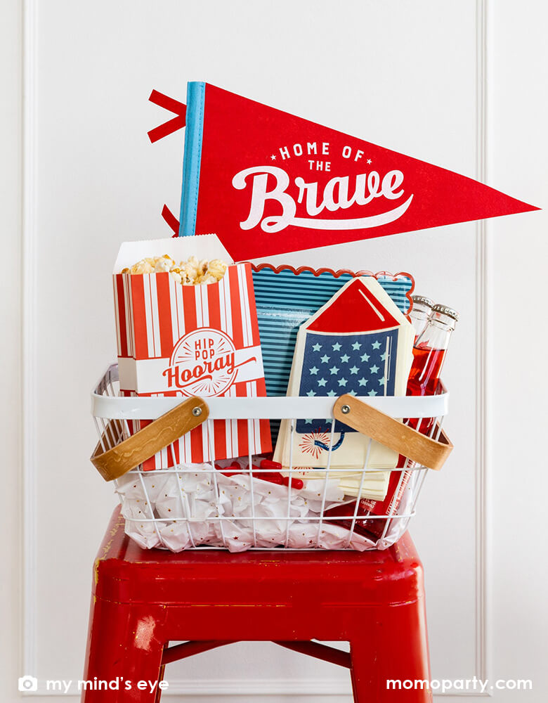 An occasion bin placed on a red stoop which is filled with festive patriotic themed party goods including Momo Party's 15" Home of the Brave red felt pennant by My Mind's Eye, blue stripe scallop-edged plates, rocket shaped guest napkins, red striped popcorn treat boxes with "hip hip hooray" on it, these are perfect decorations for a Memorial Day celebration or 4th of July Independence Day party!